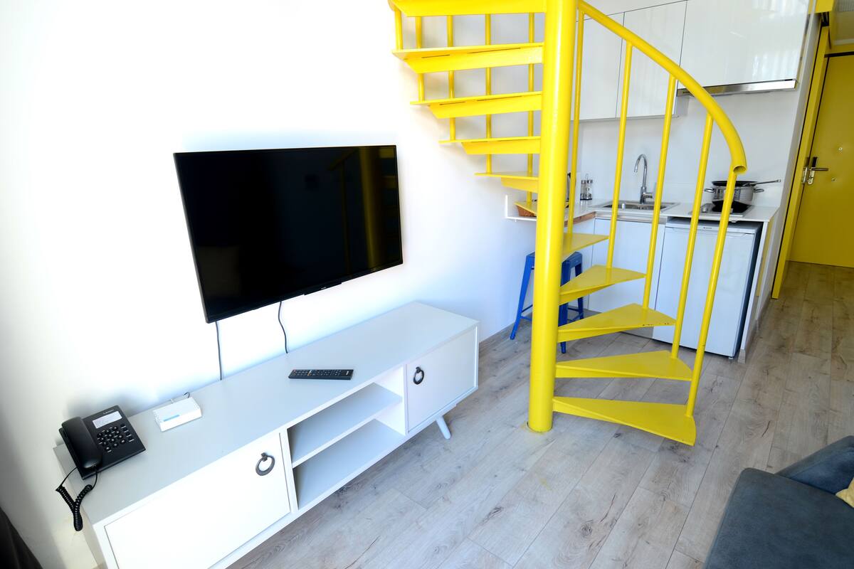 A modern living room with a flat-screen TV, a white console table, a yellow spiral staircase, and a kitchen area in the background.