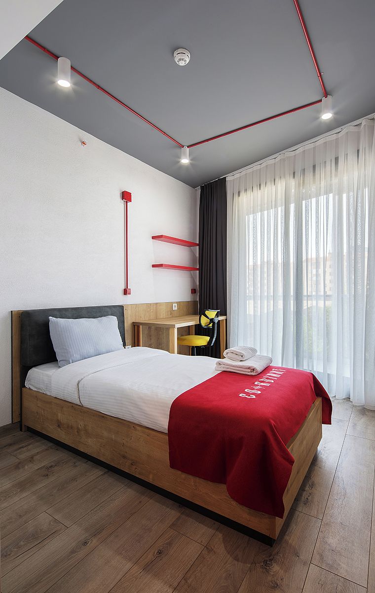 Een moderne hotelkamer met een houten bed, een rode accentmuur, een bureau met een stoel en een groot raam met dunne gordijnen.