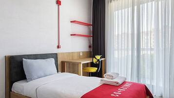 Een moderne hotelkamer met een houten bed, een rode accentmuur, een bureau met een stoel en een groot raam met dunne gordijnen.