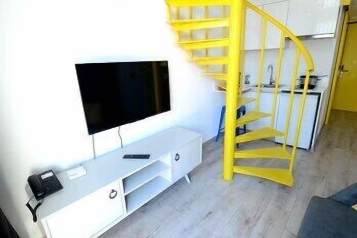 A compact living space with a TV mounted on a white cabinet, a yellow spiral staircase, and a kitchenette area.