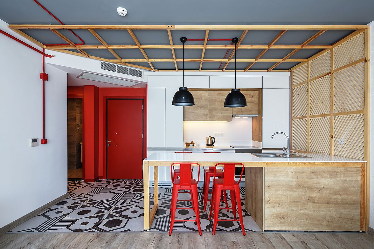 Une cuisine moderne avec un plafond en bois, un îlot central doté de tabourets et une porte rouge en arrière-plan.