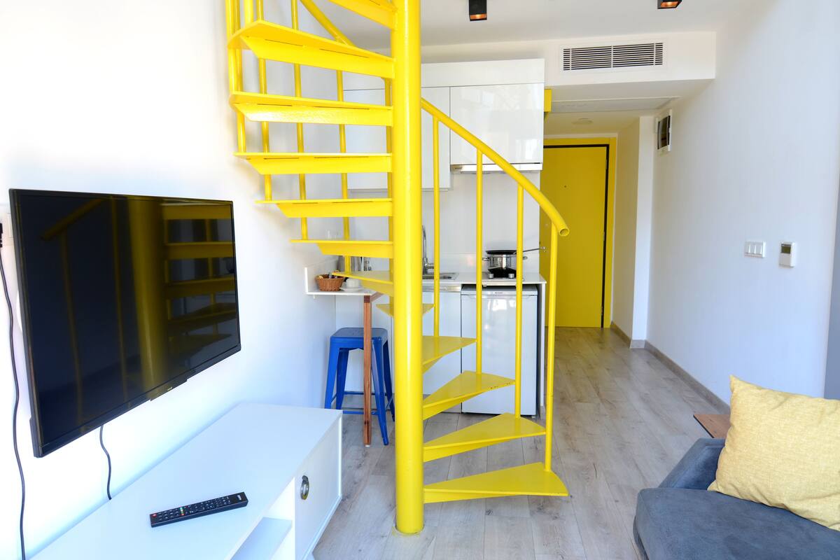A modern apartment with a yellow spiral staircase, a kitchen area, a living room with a TV, and a sofa.