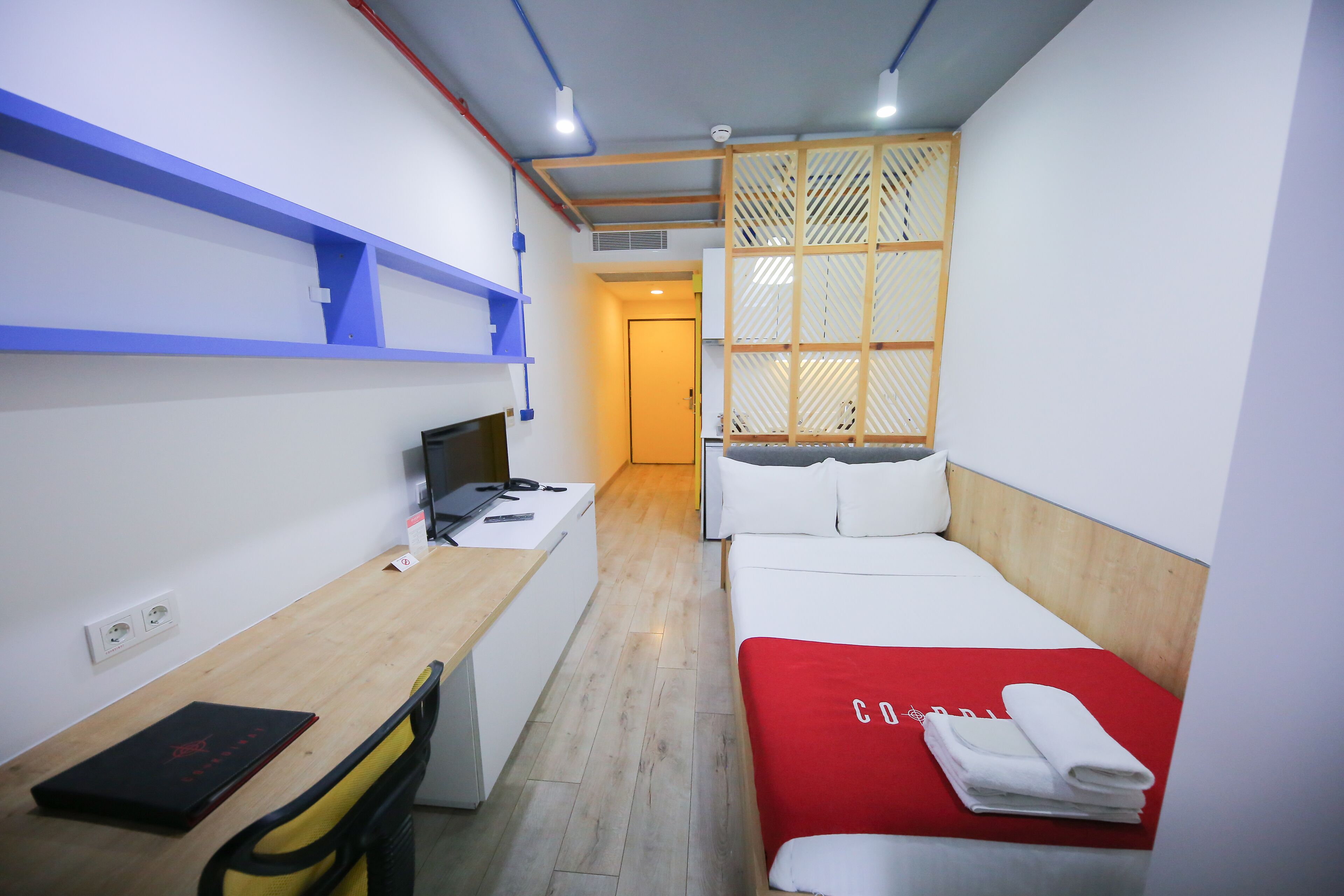 Una habitación de hotel compacta con un escritorio de madera, una cama con una colcha roja y una separador con diseño de celosía.