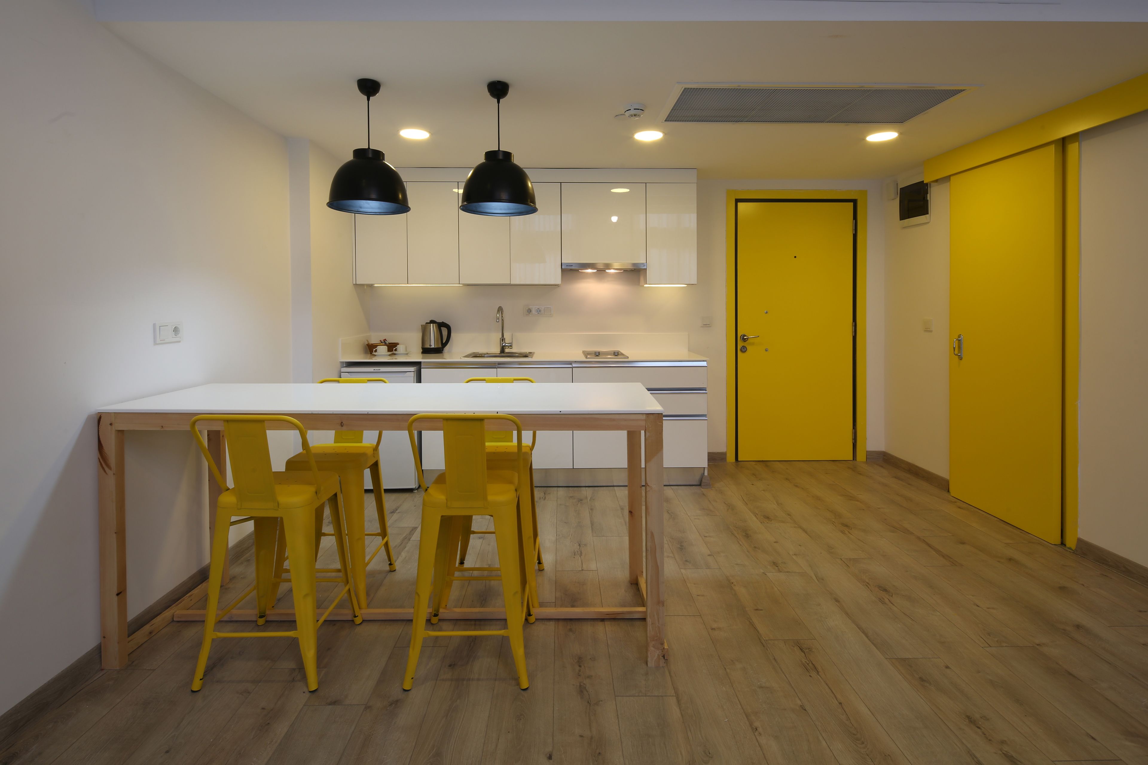 A compact kitchen with a white countertop, two yellow chairs, and black pendant lights.