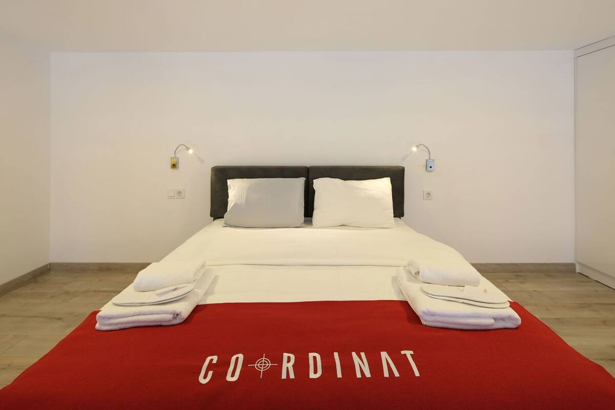 A hotel room with a bed, two bedside lamps, a red carpet with 'COORDINAT' text, and a white towel on the floor.