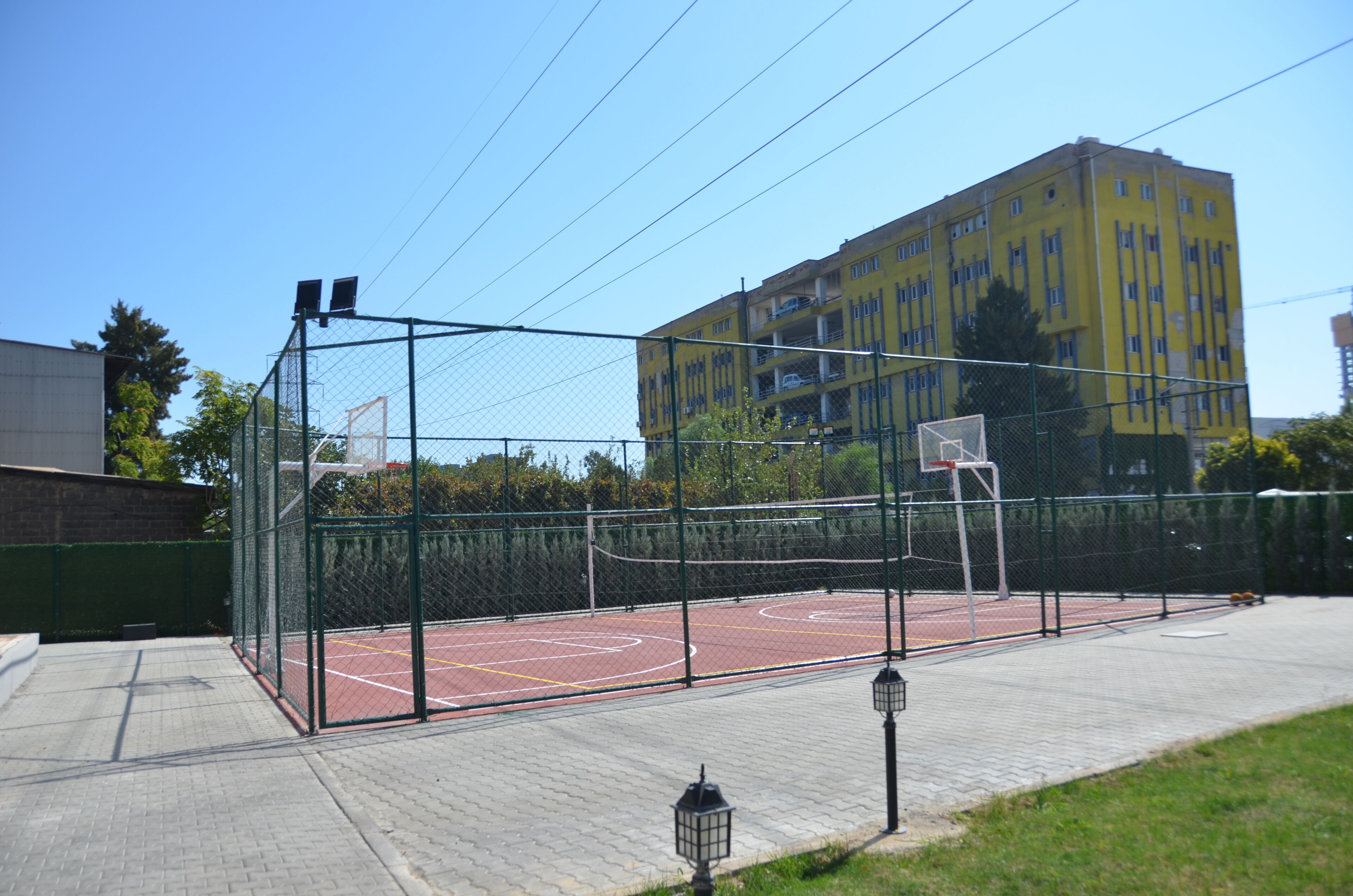 An outdoor basketball court with a chain-link fence, basketball hoops, and a paved walkway.
