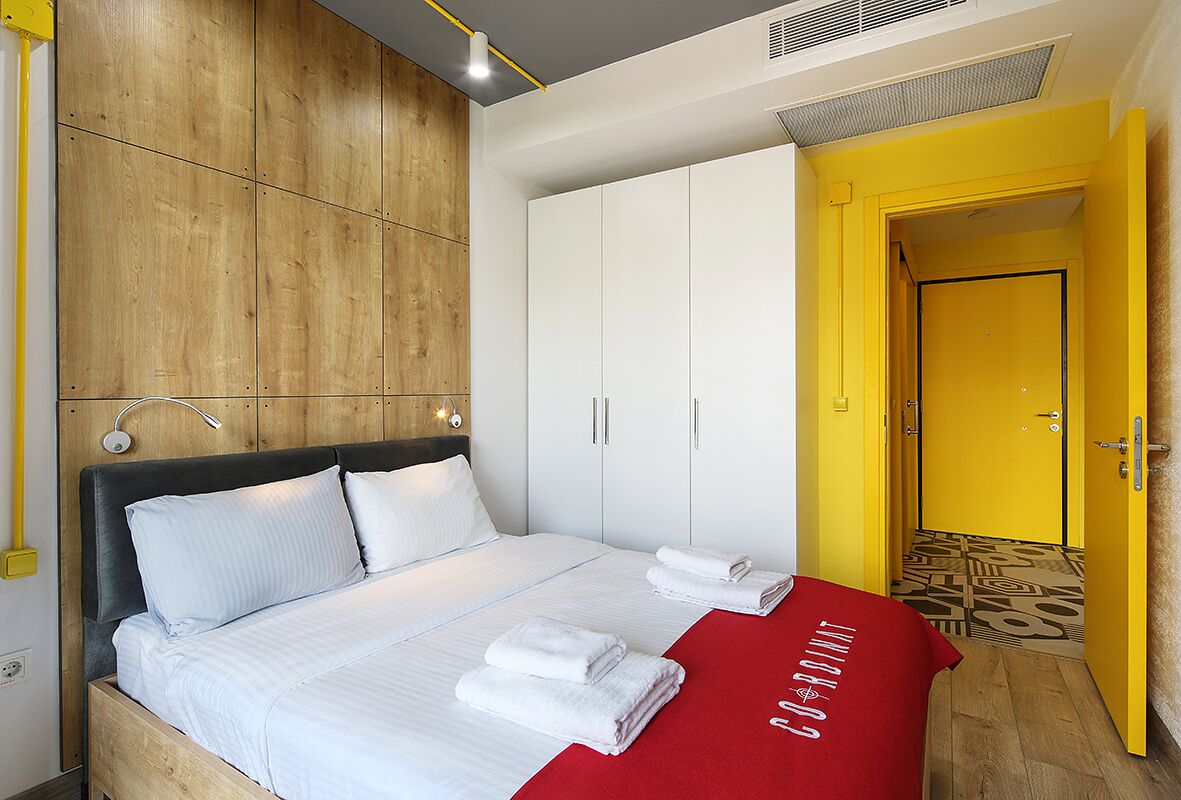 Een slaapkamer met een houten hoofdbord, wit beddengoed, een rode plaid en een gele deur.
