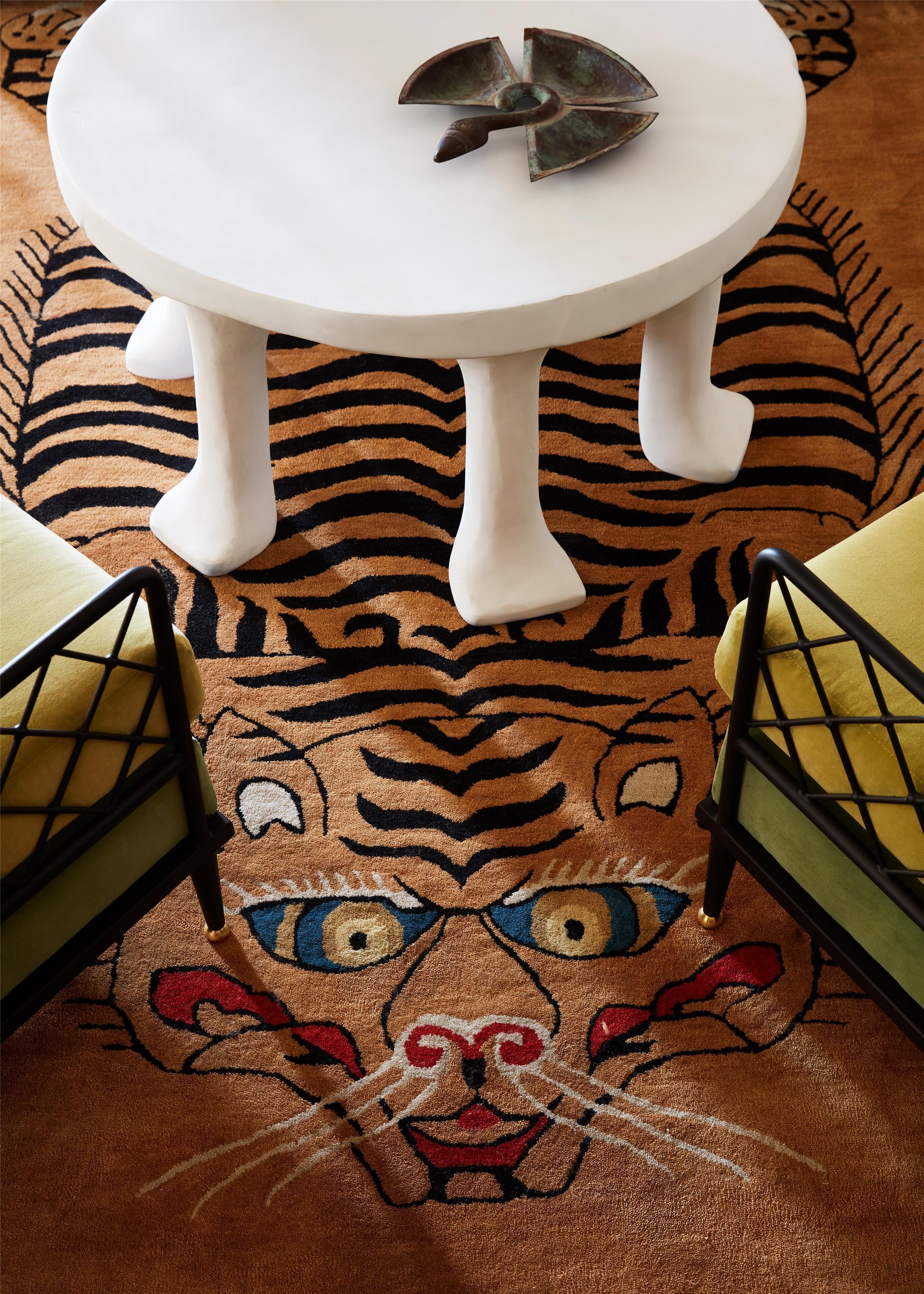Un tavolo bianco con un elemento decorativo in bronzo, un tappeto a motivi di tigre e due sedie con cuscini gialli.
