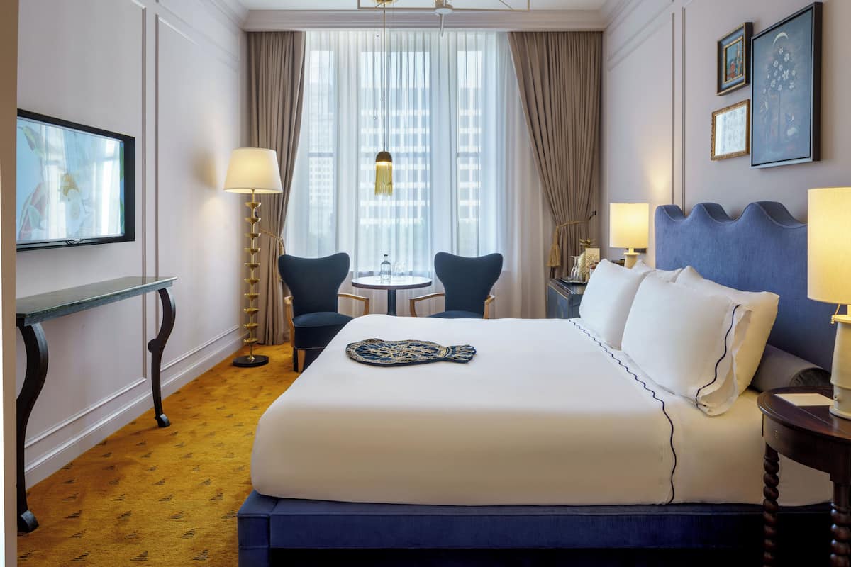 A hotel room with a large bed, a small table with chairs, a TV, and a view of the city through large windows.