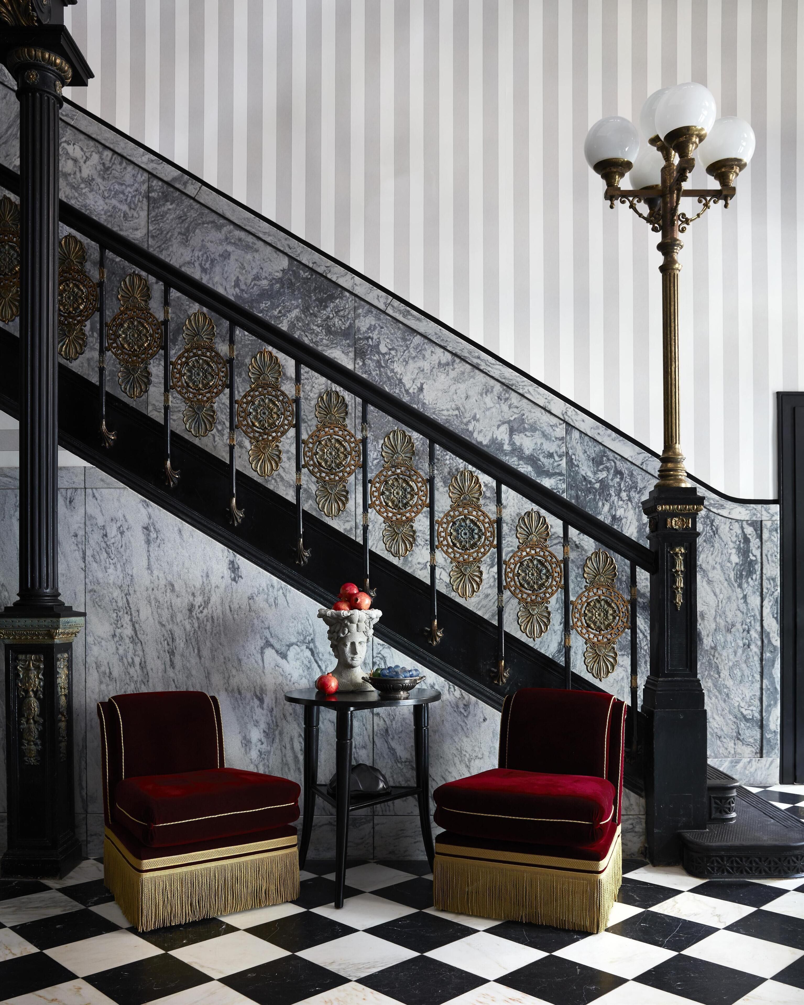 A checkered floor, a staircase with ornate railings, and two red upholstered chairs.
