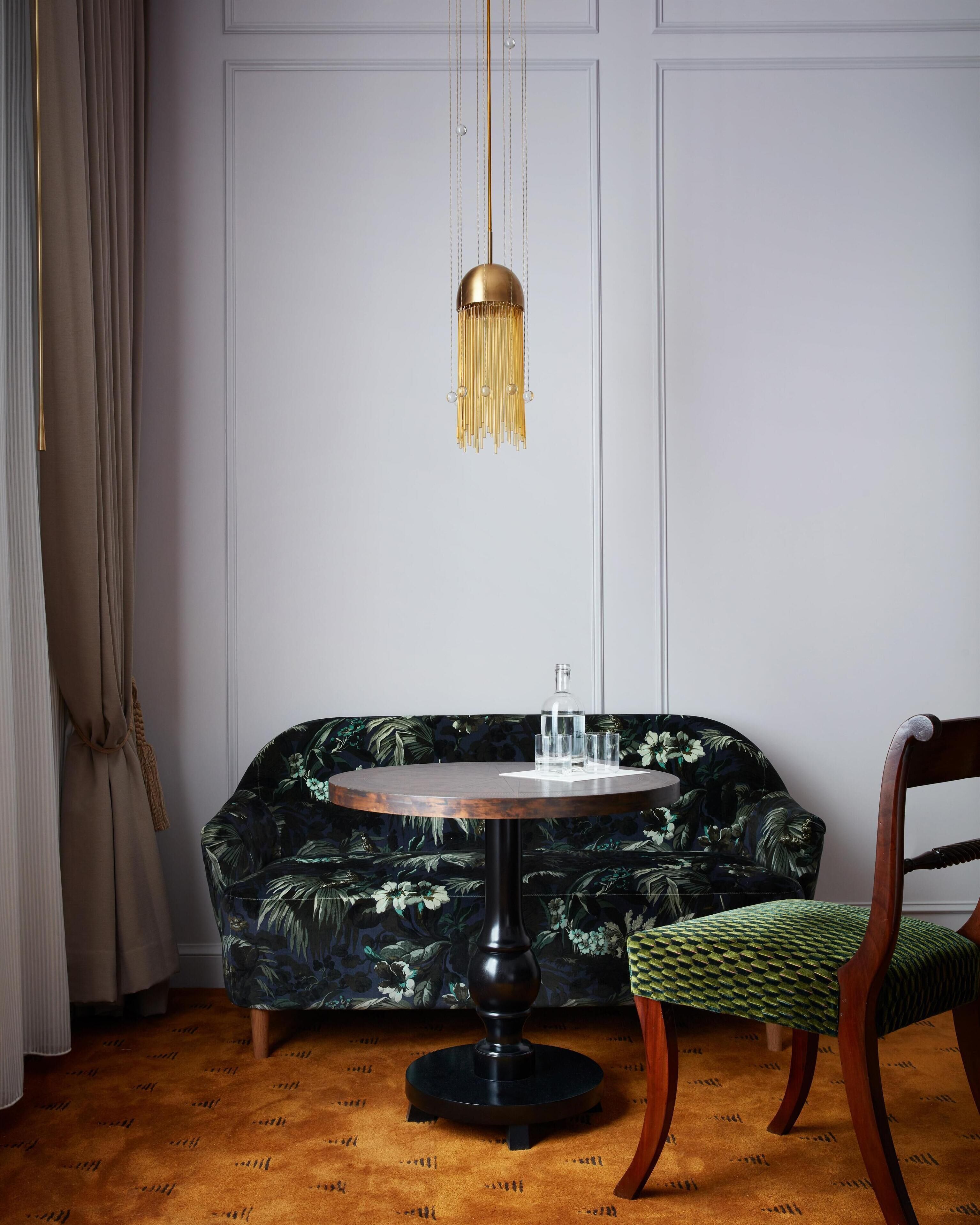 A room with a round wooden table, a green patterned sofa, a green woven chair, and a hanging light fixture.