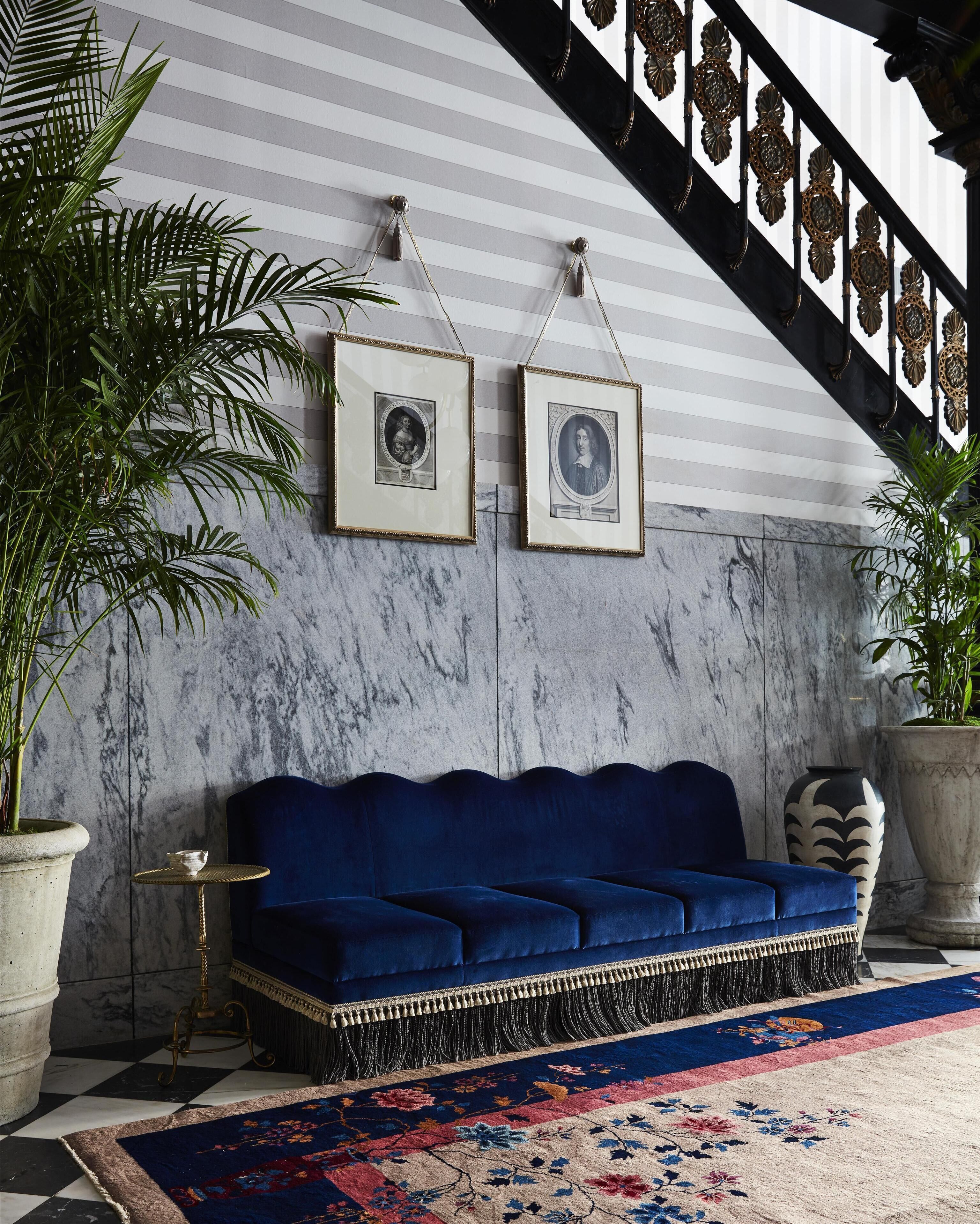 Une pièce avec un canapé bleu, deux portraits encadrés, un mur en marbre, un escalier avec des rampes décoratives et des plantes en pot.