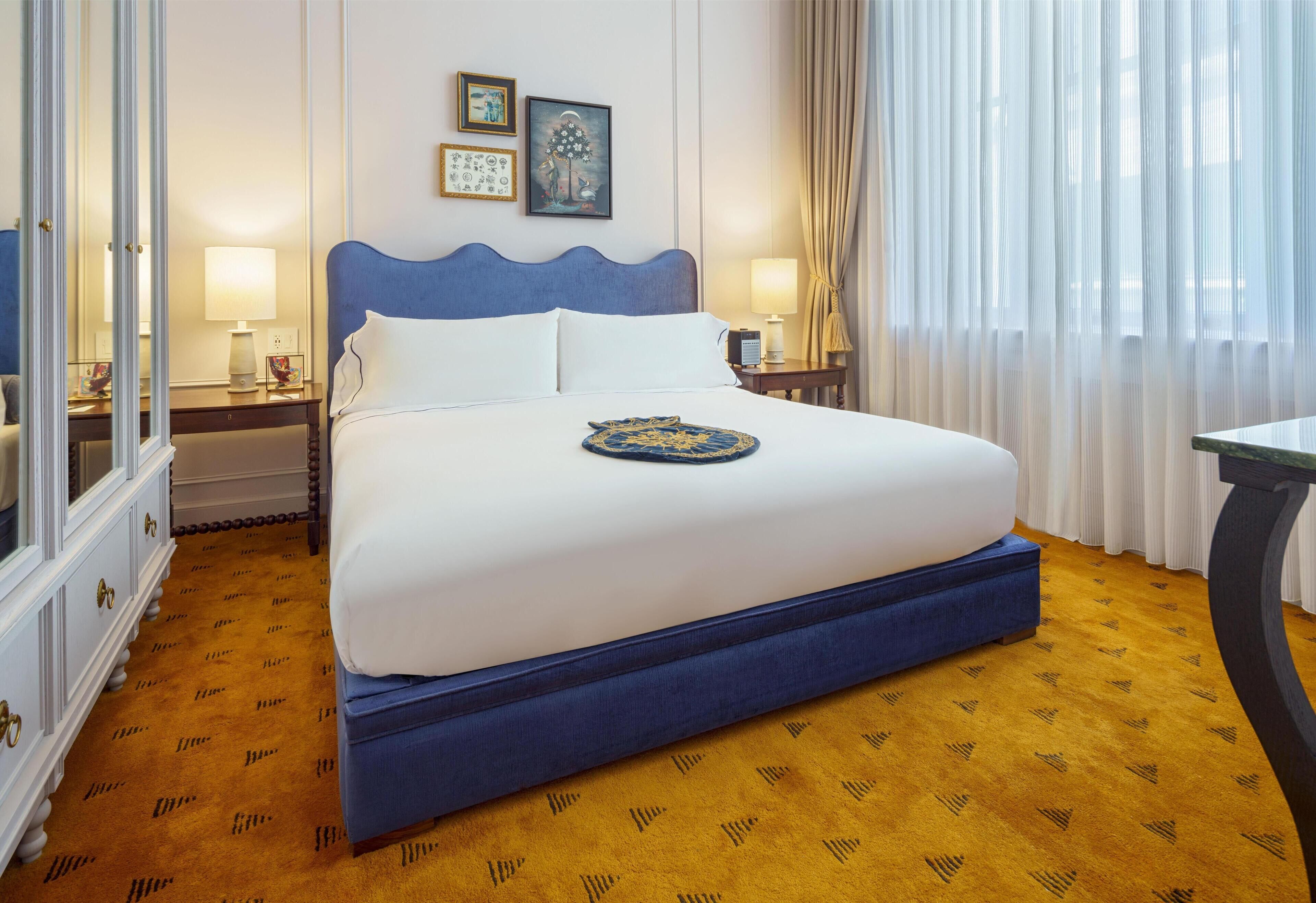 A neatly made bed with a blue headboard, white bedding, and a decorative plate on the bed. There are two bedside tables with lamps, a large mirror, and a window with sheer curtains.