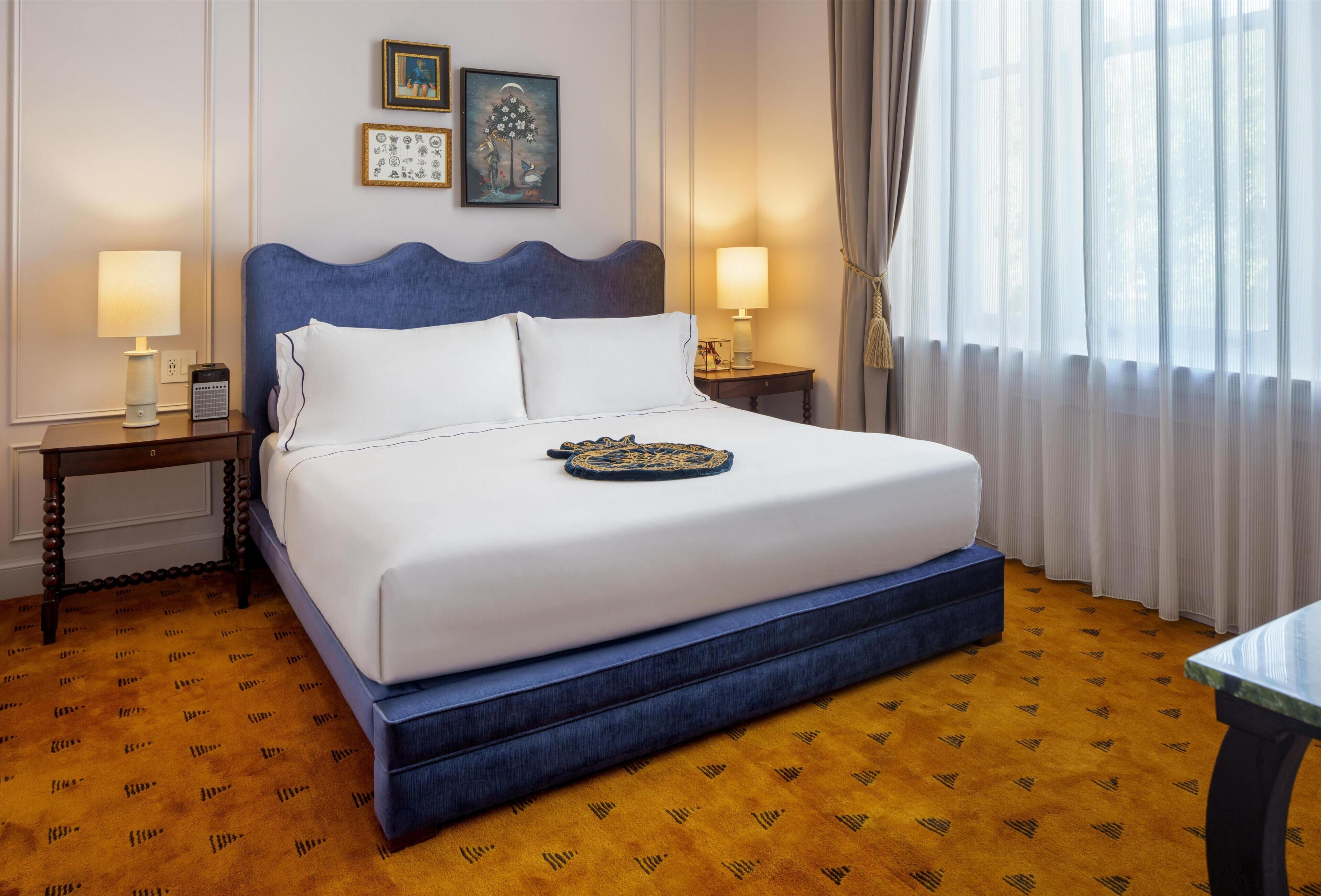 A neatly made bed with a blue headboard, two bedside tables with lamps, a framed picture on the wall, and a window with curtains.