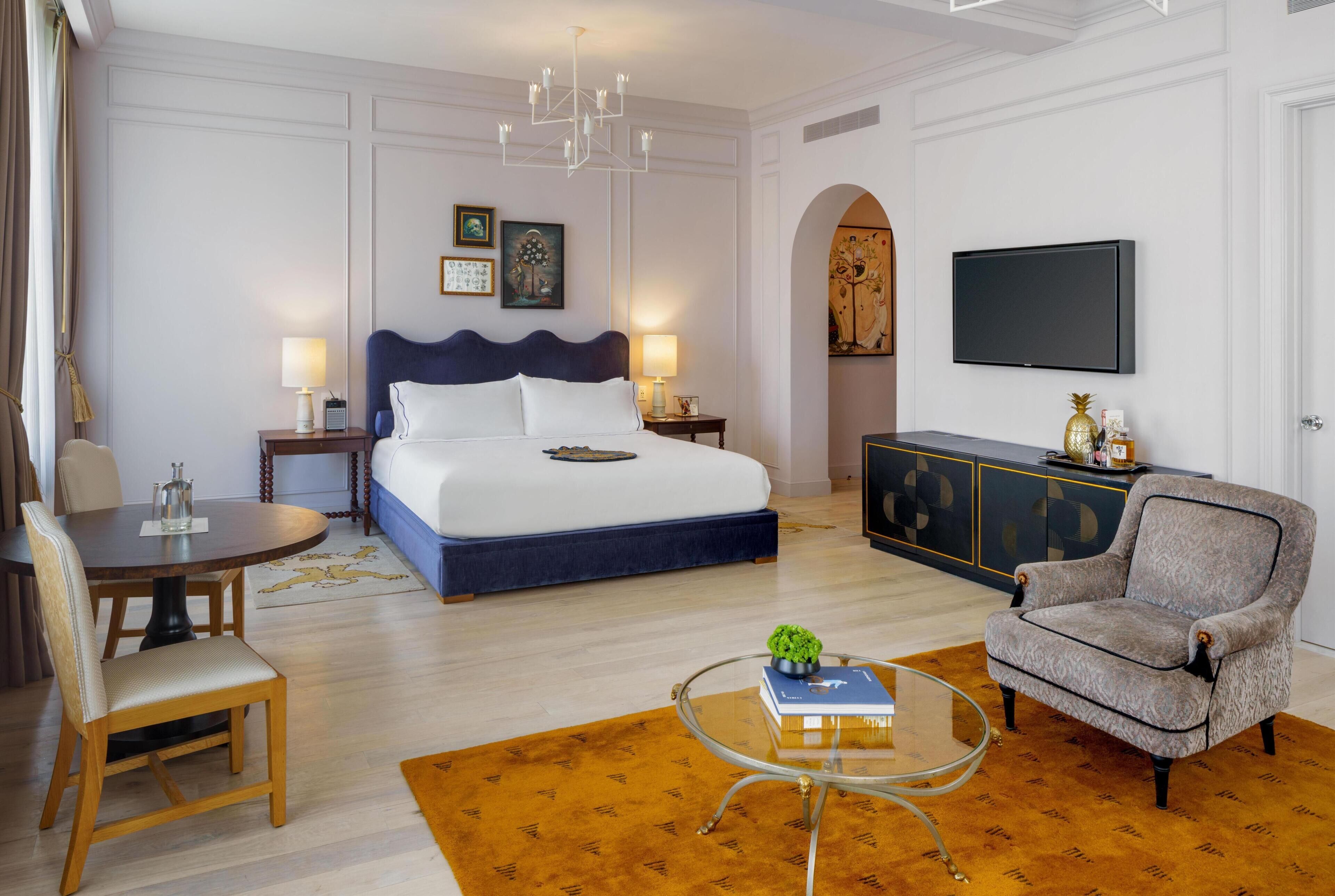 A hotel room with a large bed, a television, a dining table with chairs, and a sofa.