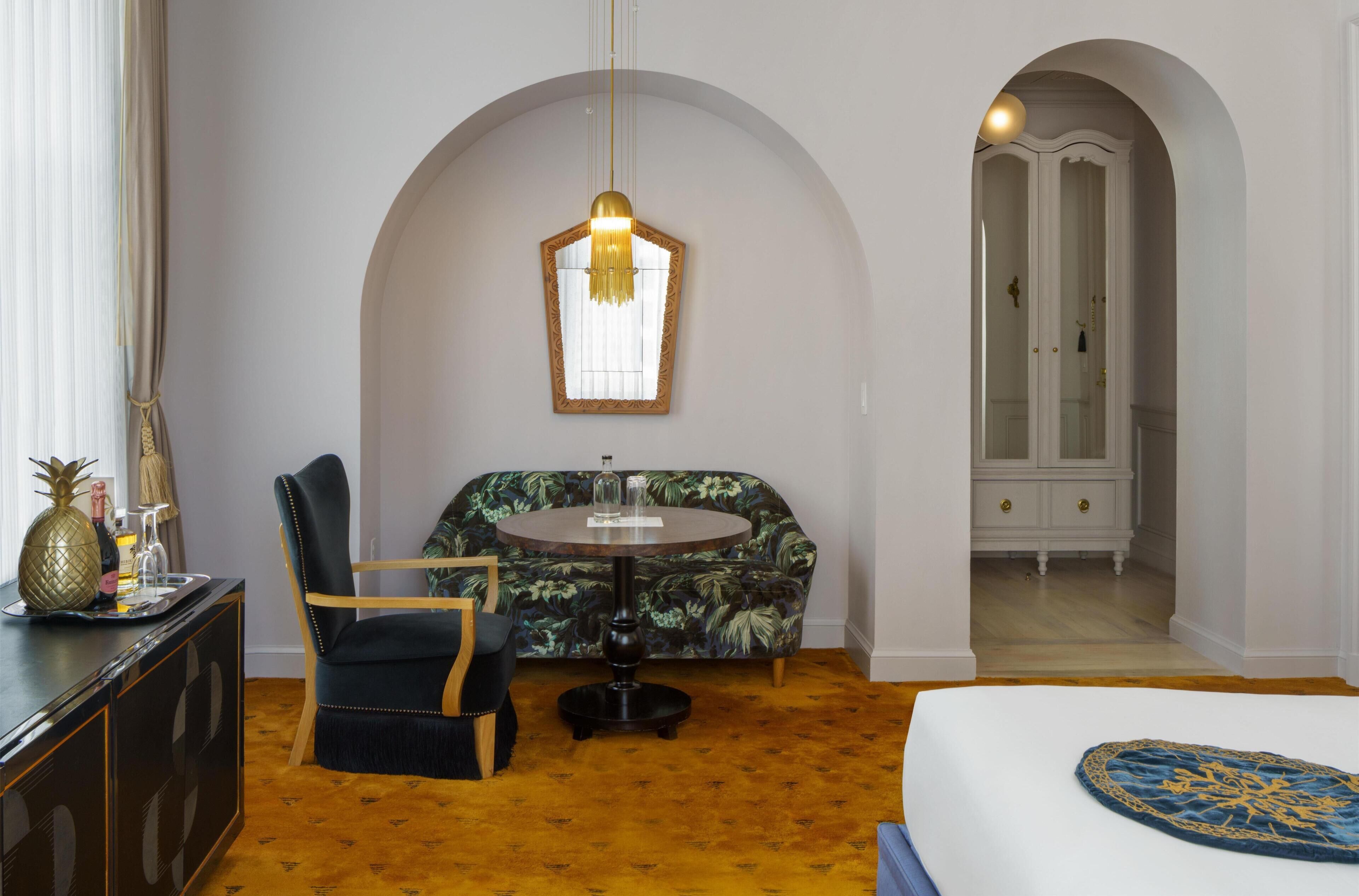 A hotel room with a round table, a sofa, a chair, a bed, and an arched doorway.