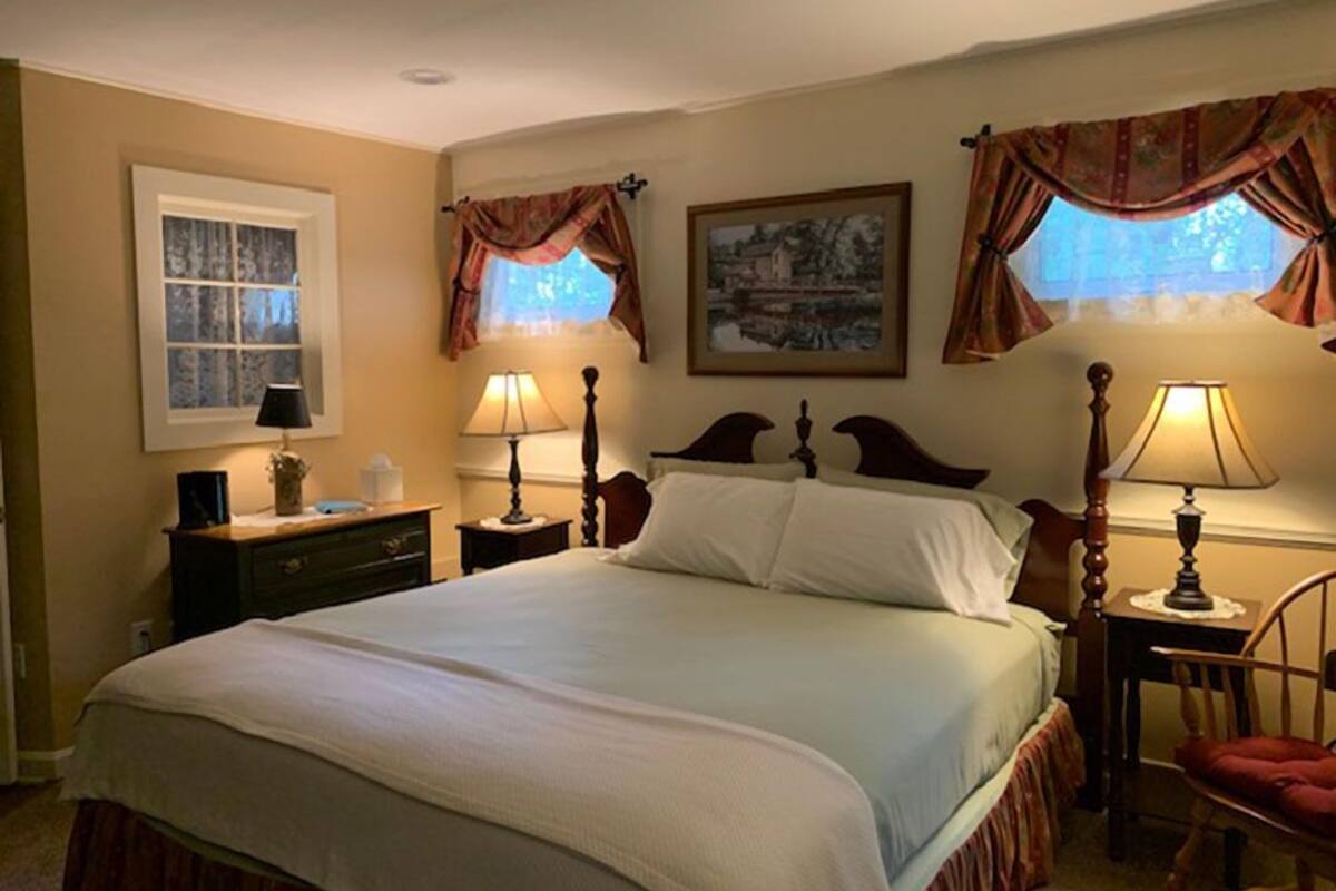 Une chambre avec un grand lit, deux tables de chevet avec des lampes, une chaise, une fenêtre avec des rideaux et un tableau encadré au mur.