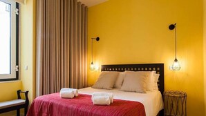 Hotellihuone, jossa on sänky, kaksi yöpöytälamppua, penkki ja ikkuna, jossa on verhot.