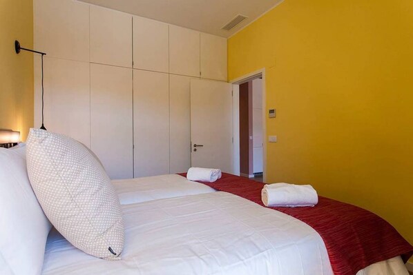 Habitación de hotel con dos camas, una colcha roja y blanca, un almohadón blanco y un cabecero blanco.