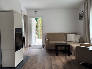 Ein modernes Wohnzimmer mit einer Couch, einem Couchtisch und einem Fernseher. Eine Tür führt in einen Außenbereich mit Blick auf Bäume.