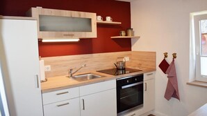 A compact kitchen with white cabinets, a double sink, a stove, and a microwave. There are wooden shelves above the sink and a window to the right. - Haus Hanna (Dagebuell)