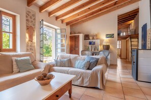A cozy living room with a sofa, a coffee table, and a kitchen area with a refrigerator and shelves. - Country House with Romantic Patio and Pool – Finca Ca'n Vaqueta (Soller)