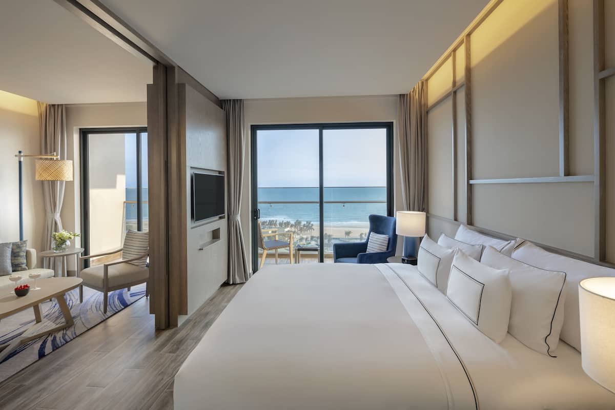Een hotelkamer met een groot bed, een bureau met stoel, een televisie en uitzicht op het strand.