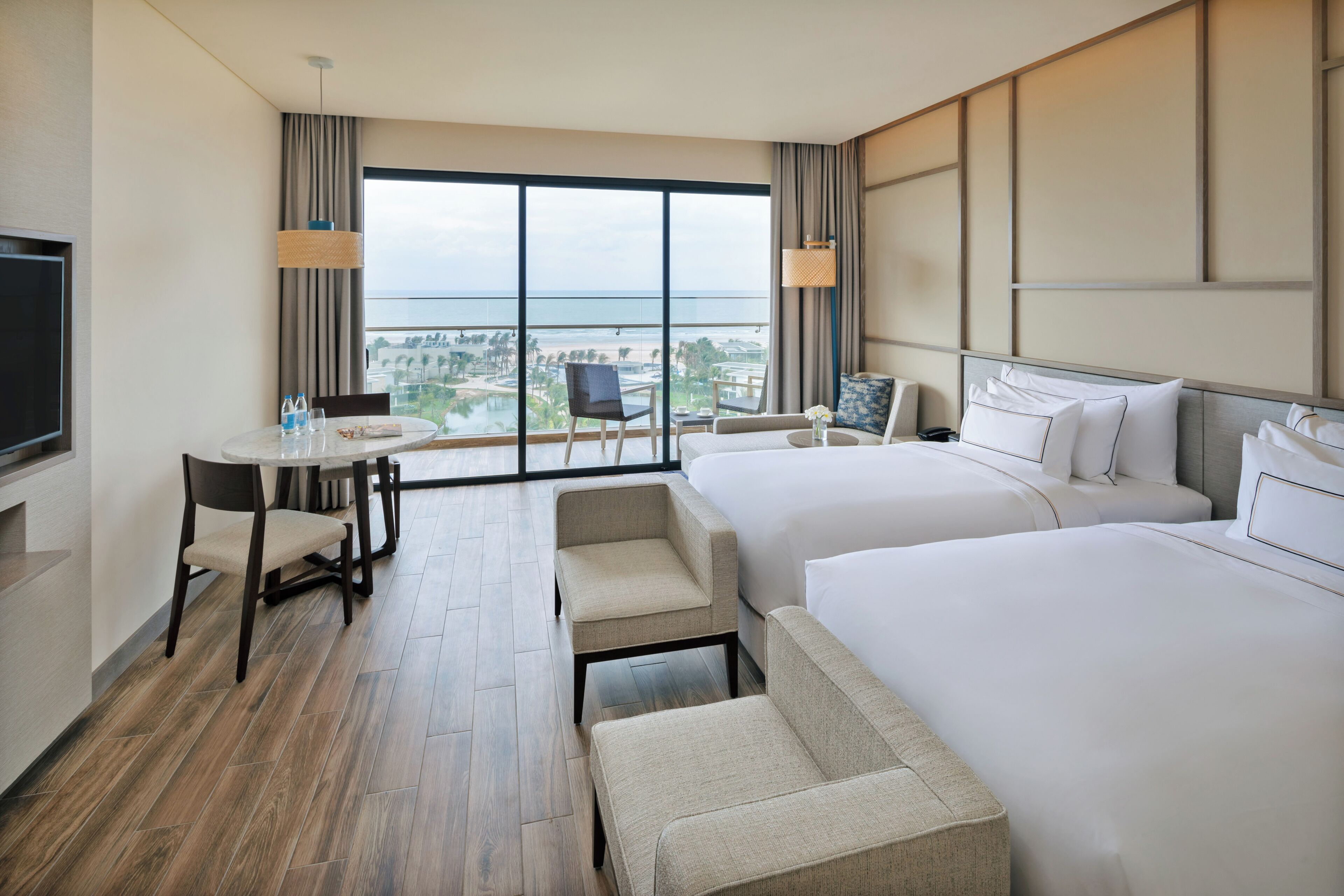 A hotel room with two beds, a dining table, a TV, and a view of the city and ocean.