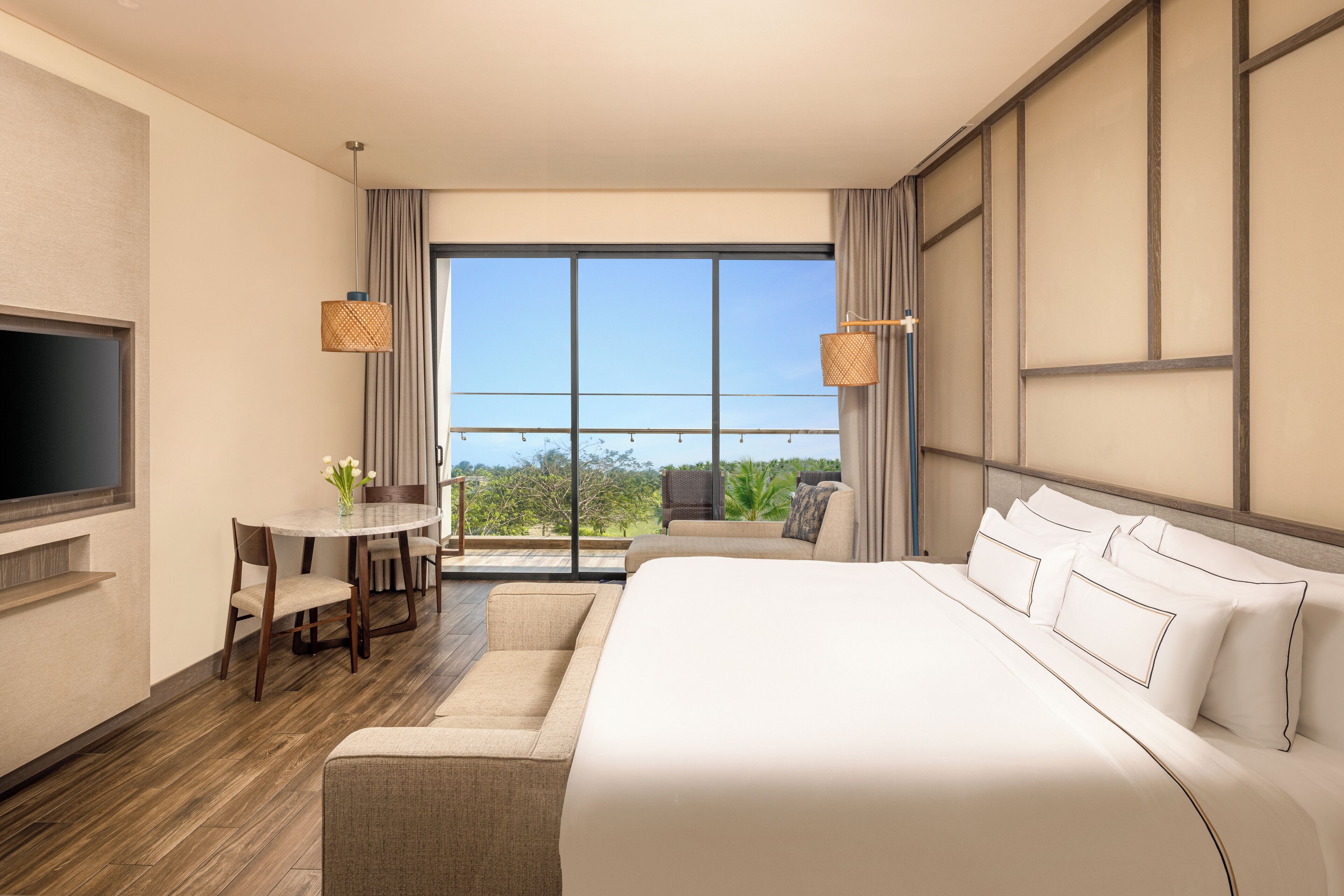 Una habitación de hotel moderna con una cama grande, un televisor de pantalla plana, una zona de comedor con mesa y sillas, y un ventanal con vistas al exterior.
