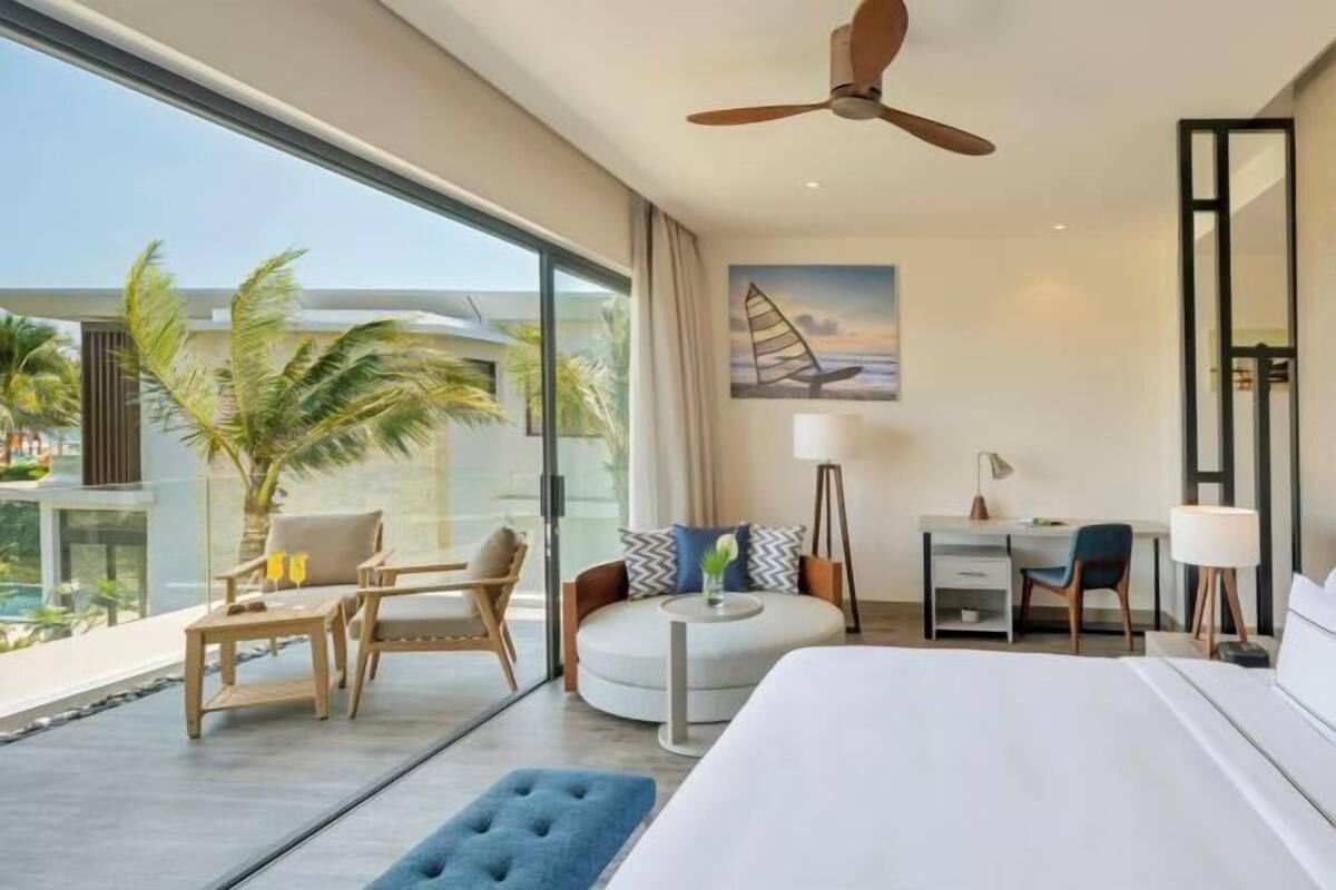 Een moderne hotelkamer met een groot bed, een bureau, een stoel, een ronde zithoek en uitzicht op palmbomen en een zwembad.