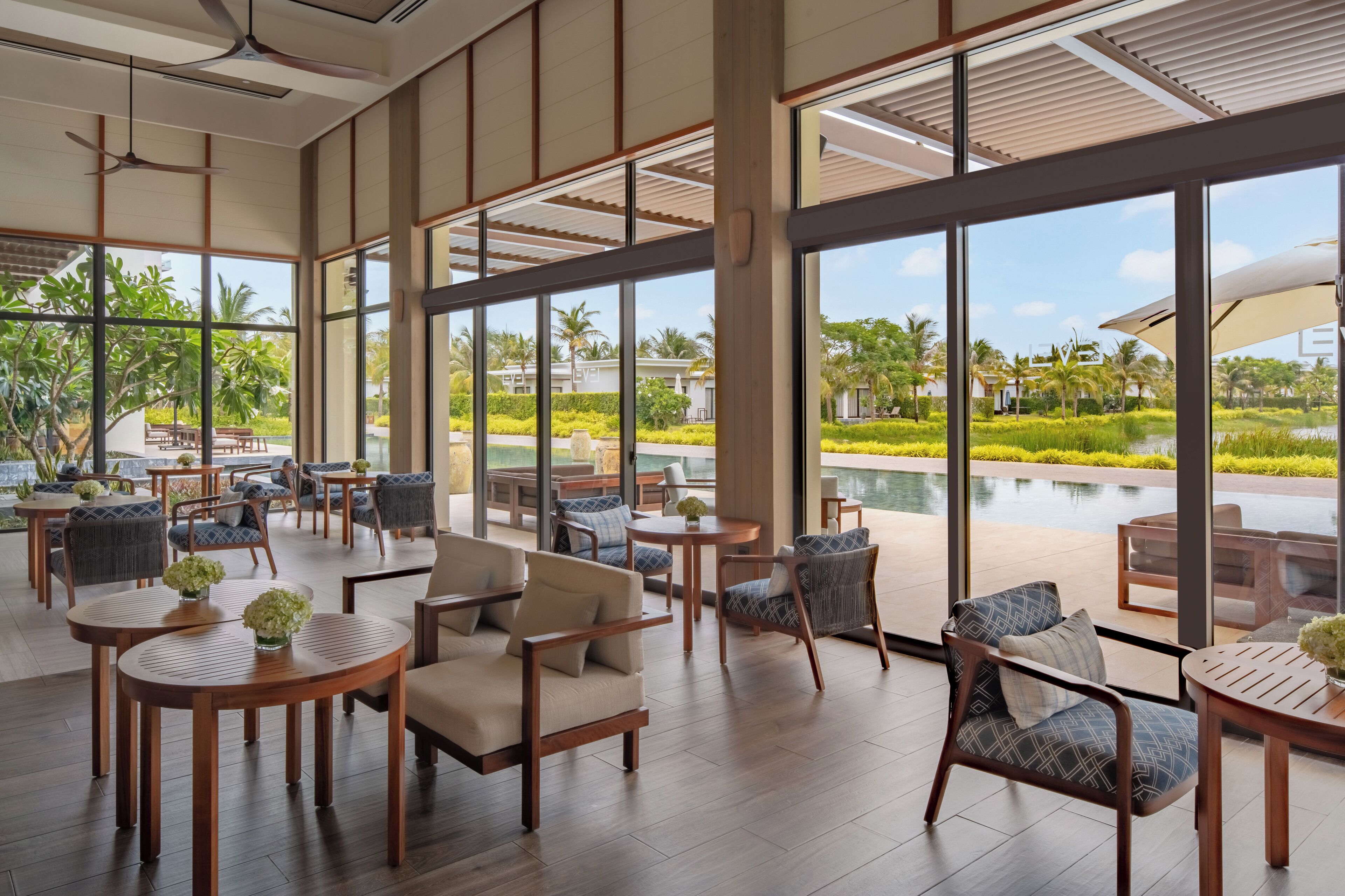 A spacious indoor area with large windows, wooden furniture, and a view of a pool and greenery.