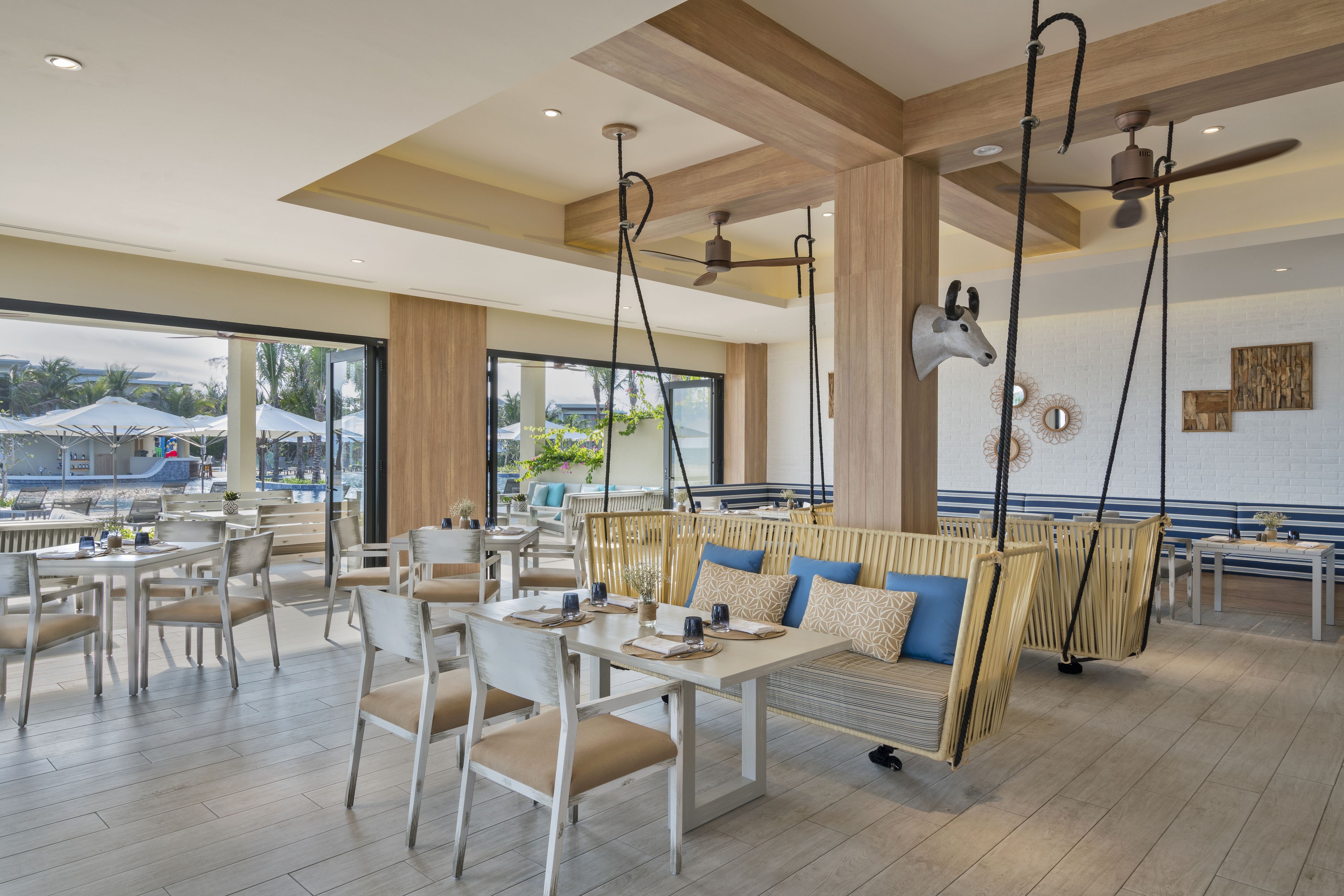 A spacious outdoor dining area with white tables and chairs, a hanging swing chair, and a ceiling fan.