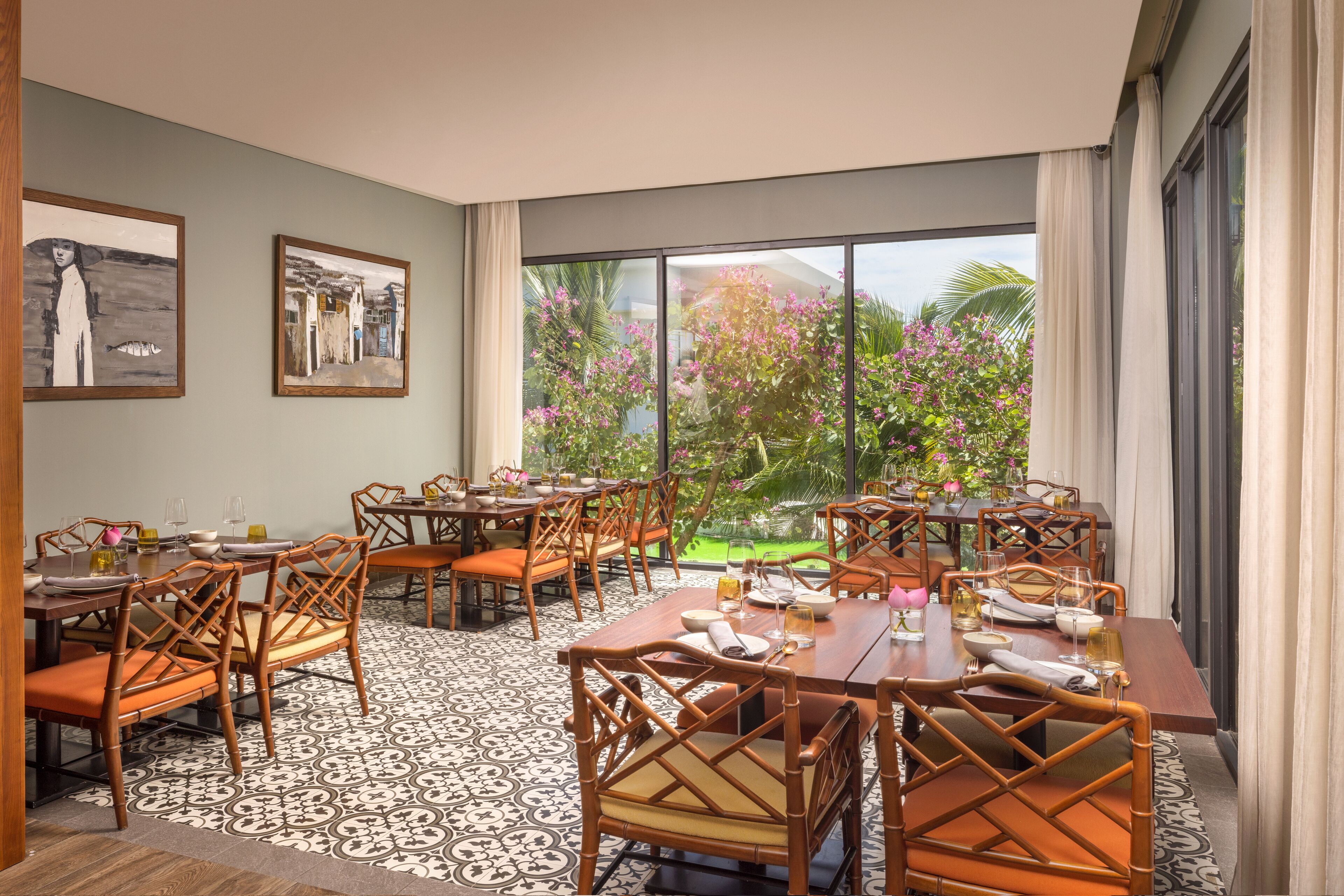 Un comedor con varias mesas y sillas, ventanas amplias con vistas a un jardín y obras de arte en las paredes.