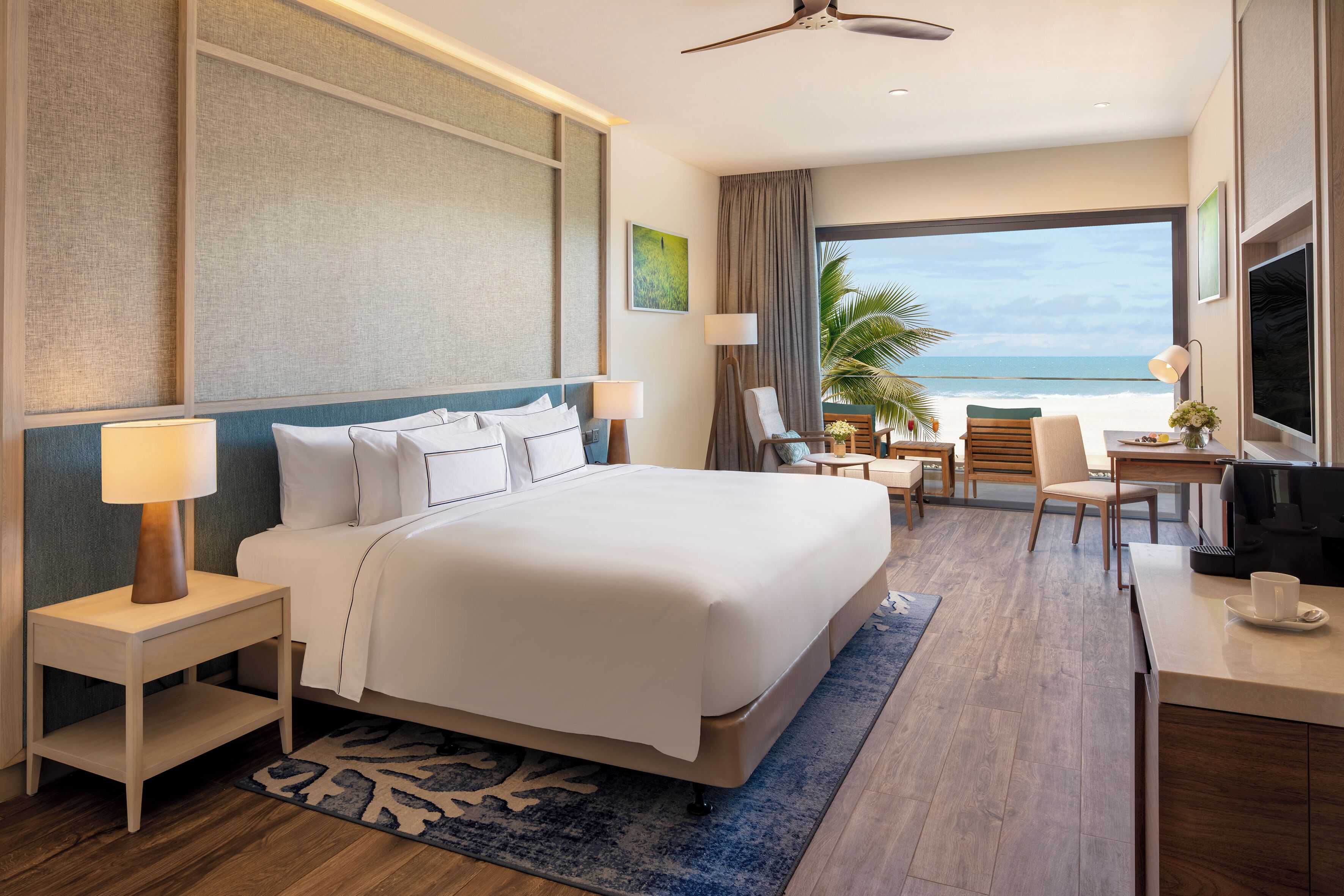 A hotel room with a large bed, a desk, a chair, a TV, and a view of the ocean.