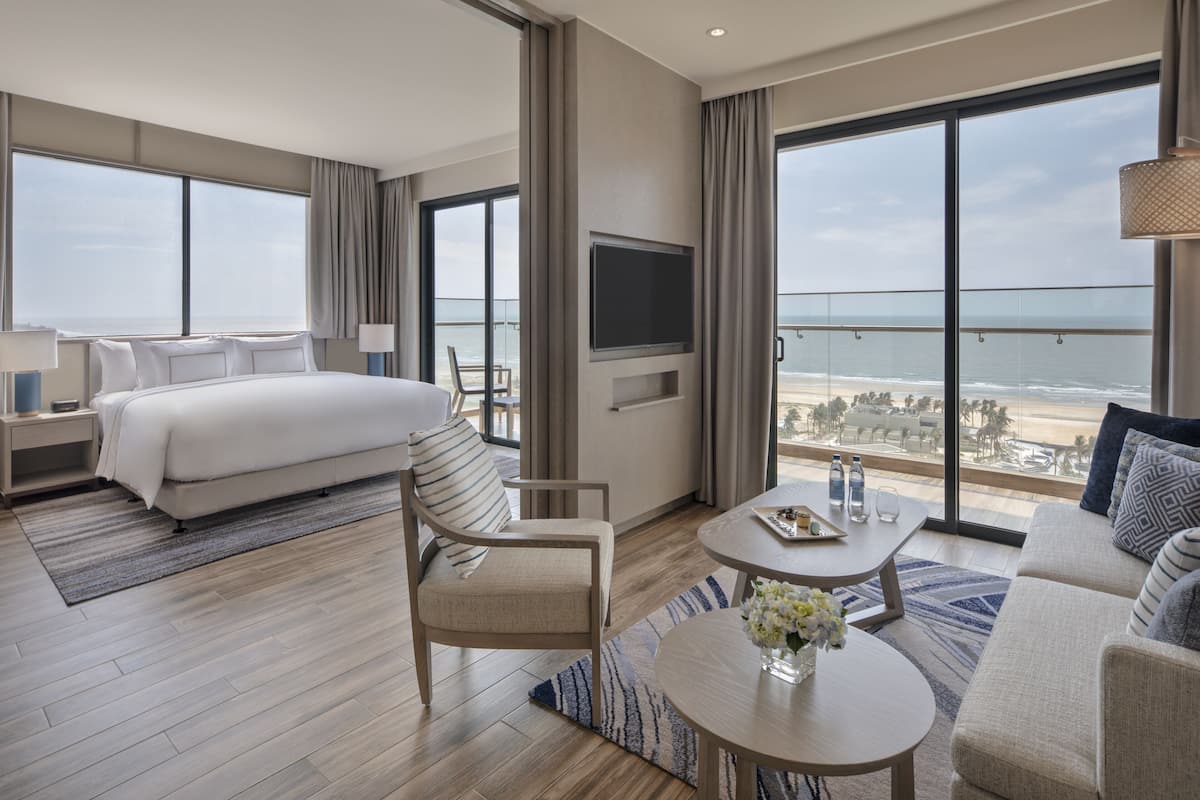 Een moderne hotelkamer met een groot bed, een zithoek met een bank en fauteuils, een eettafel en een aan de muur bevestigde televisie.