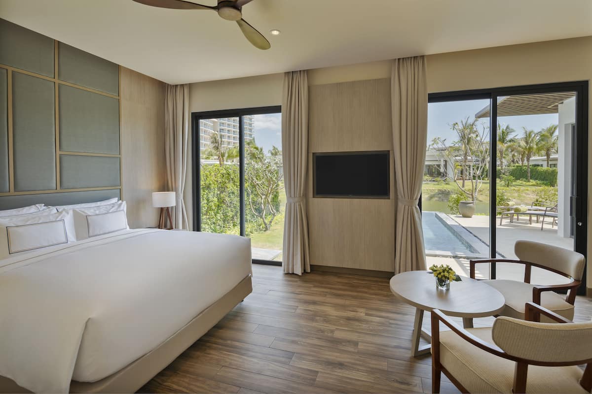 Een moderne hotelkamer met een groot bed, een flatscreen televisie, een eettafel en uitzicht op een zwembad en palmbomen.