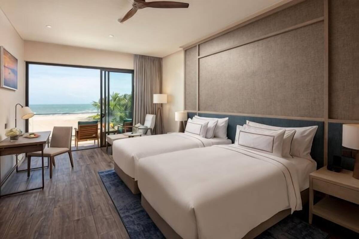 Hotelkamer met twee bedden, een bureau, een stoel, een lamp en uitzicht op het strand.