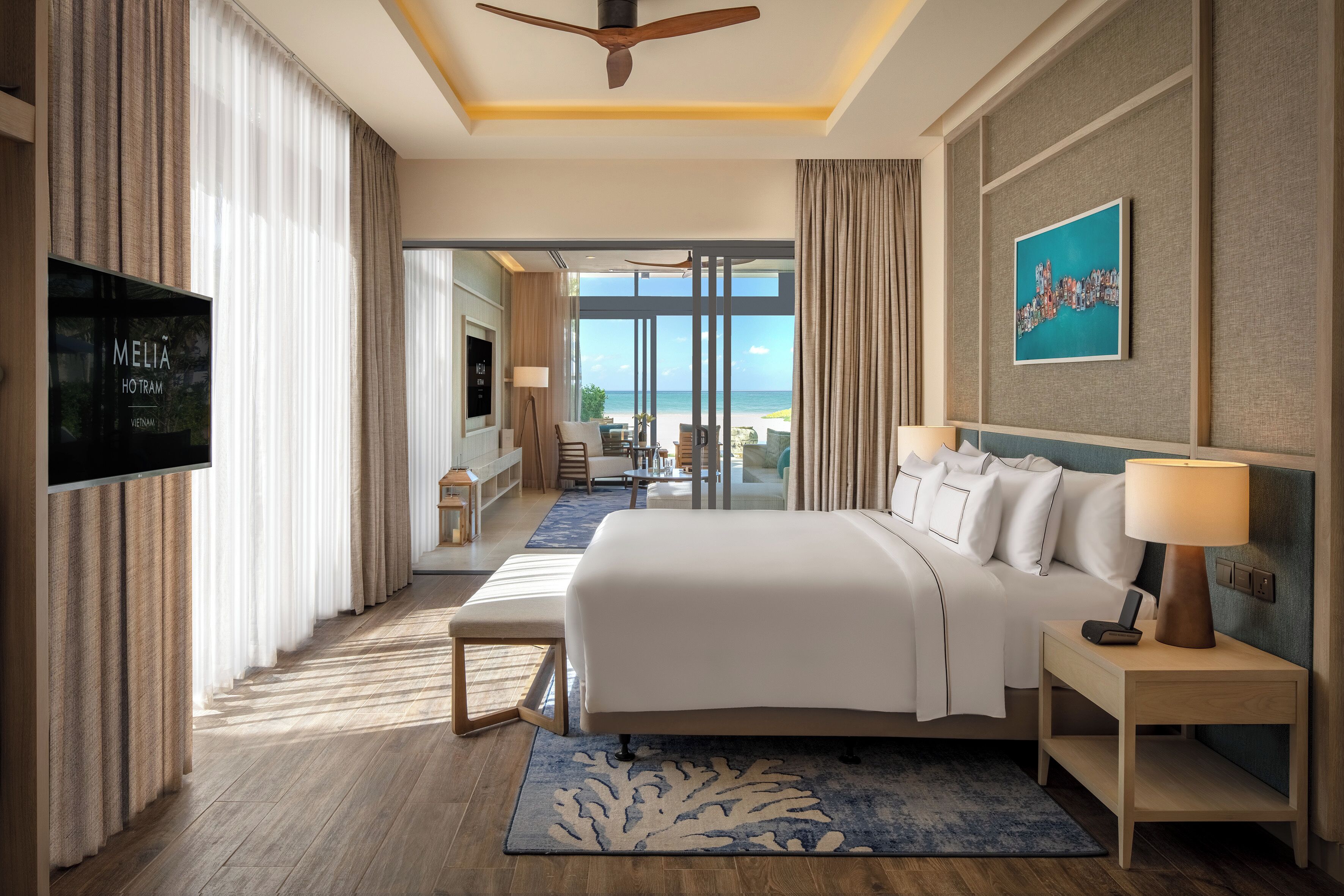A hotel room with a bed, bedside table, TV, and a view of the beach.