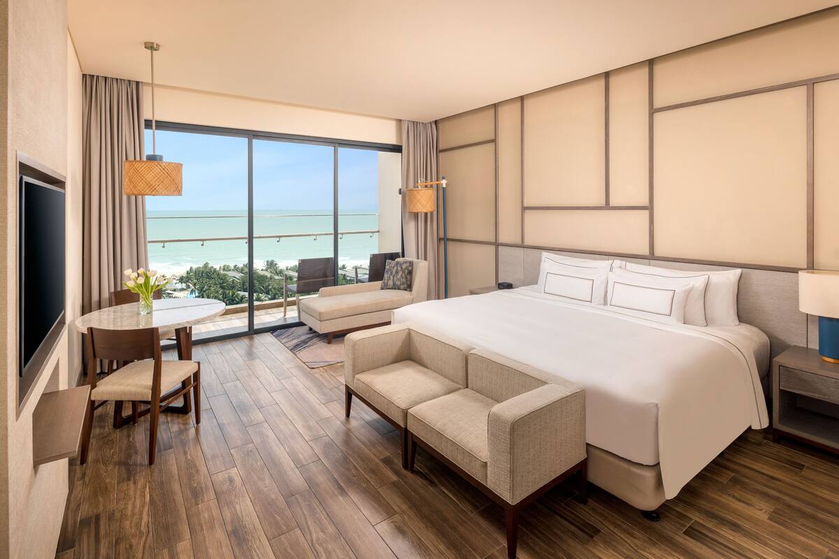 Een hotelkamer met een groot bed, een bank, een kleine tafel met een vaas en uitzicht op de oceaan.