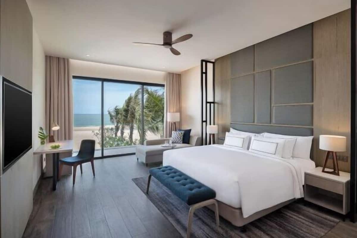 Een moderne hotelkamer met een groot bed, een bureau, een stoel, een bankje, een plafondventilator en uitzicht op de oceaan.