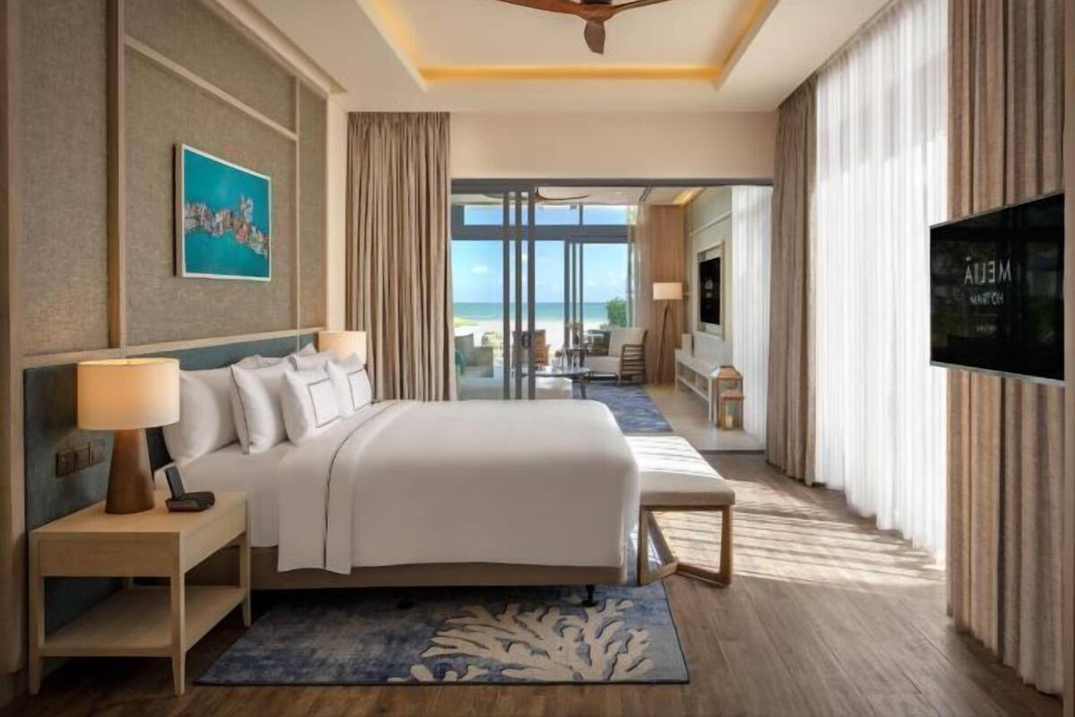 Een hotelkamer met een bed, een televisie, een bureau met lamp en een groot raam, met uitzicht op het strand.
