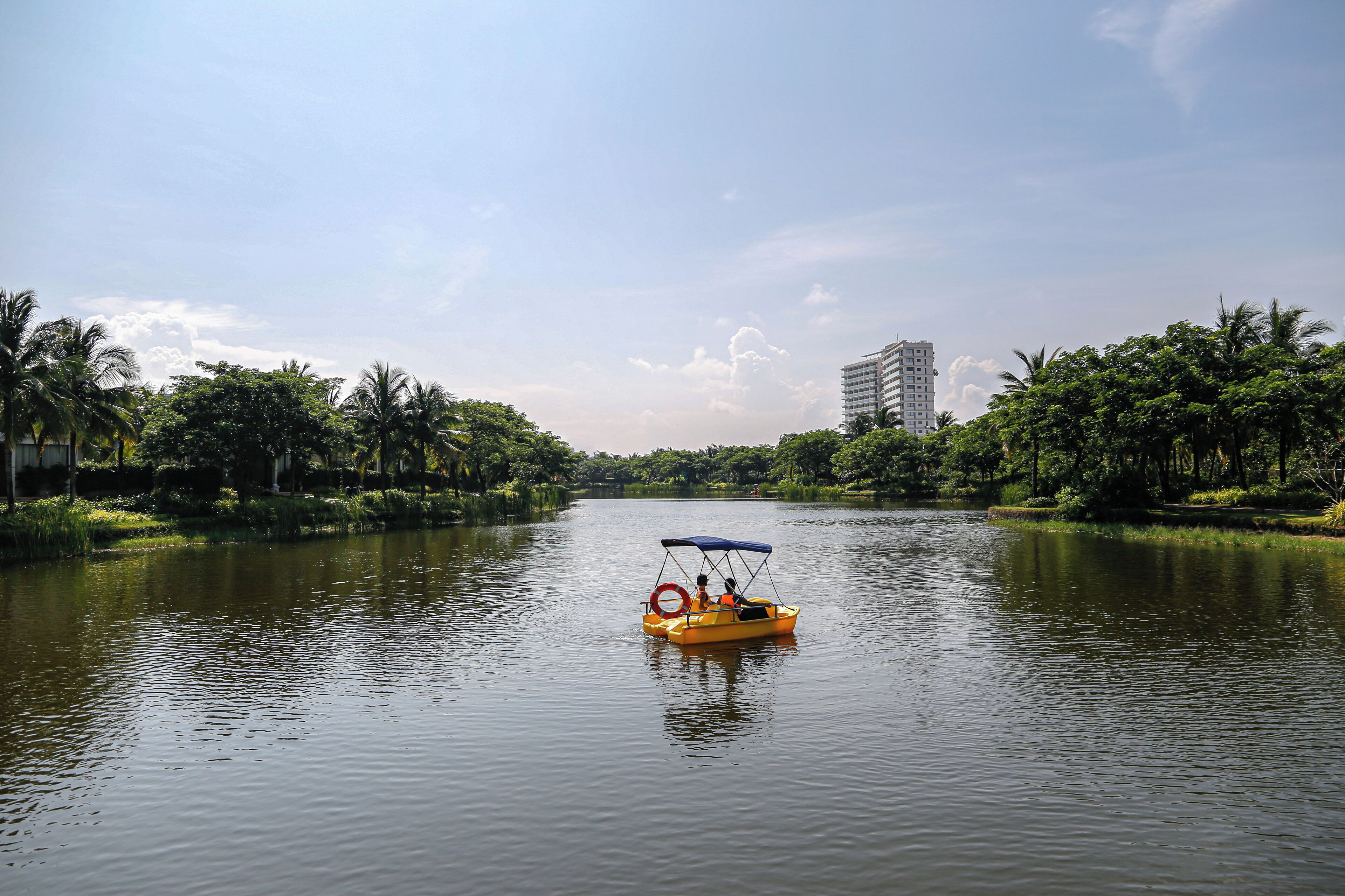 A river with a yellow boat, surrounded by greenery and a tall building in the background.