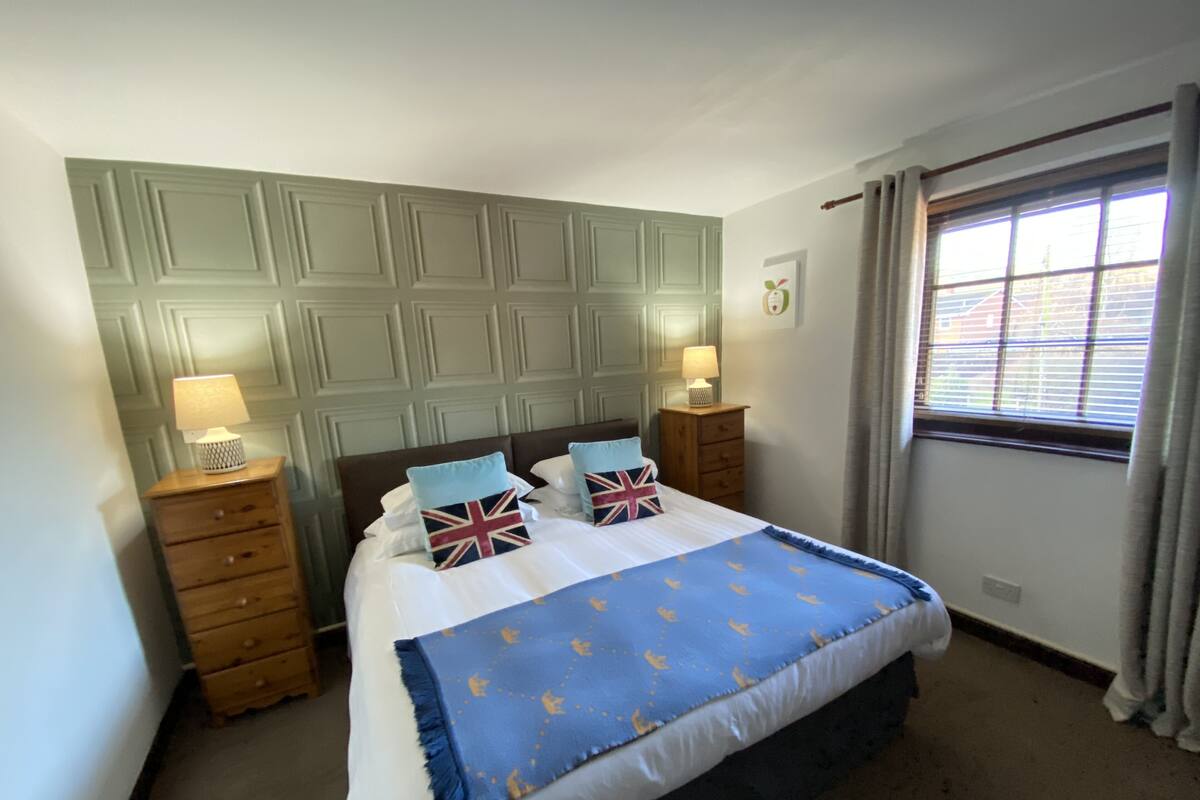 Un dormitorio ordenado con una cama, dos mesitas de noche con lámparas, una ventana con cortinas y un panel decorativo en la pared.