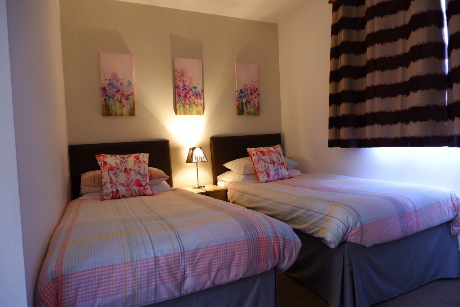 Habitación de hotel con dos camas, cada una con un cojín estampado con motivos florales y una colcha a cuadros. Hay tres cuadros colgados en la pared sobre las camas, y una cortina rayada en la ventana.