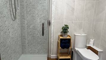 Ett modernt badrum med en duschavdelning med glasväggar, en toalett och en liten hylla i trä med handdukar och en växt.