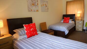 A hotel room with two beds, each with striped bedding and red pillows. There are two wall-mounted paintings of flowers, a nightstand with a lamp, a chair, and a small table with a lamp.