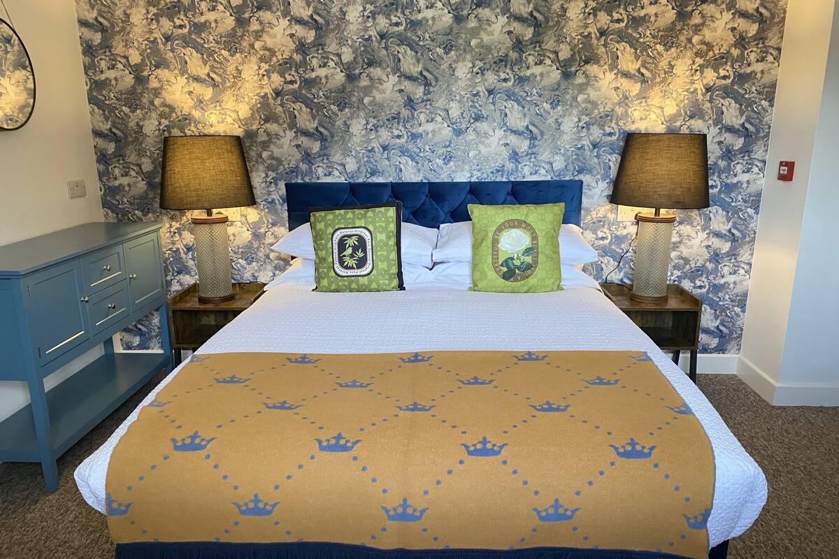 Una cama bien tendida con cabecera azul, dos mesitas de noche con lámparas, un tocador azul y un papel tapiz estampado.