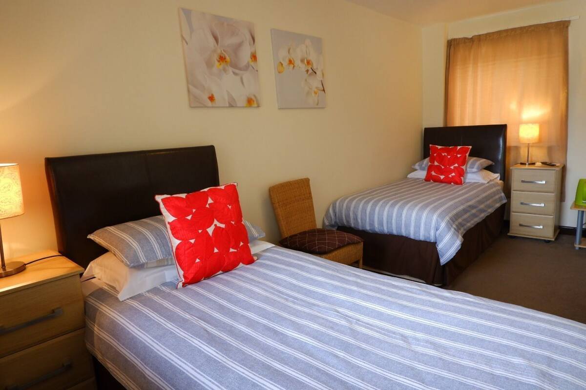 Habitación de hotel con dos camas, una mesita de noche con lámpara, una silla y una pequeña mesa con lámpara.
