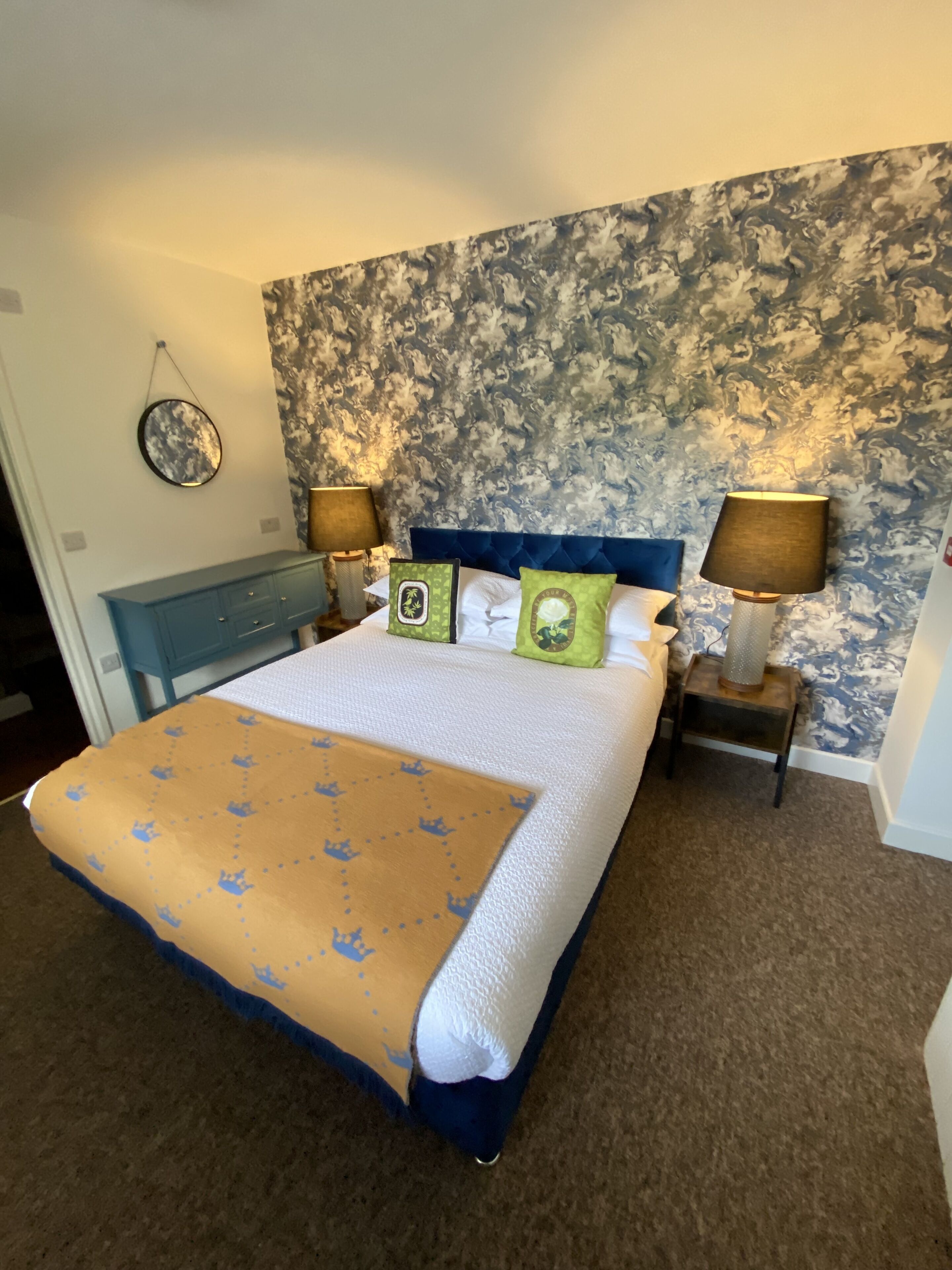 Un dormitorio con una cama grande, dos mesitas de noche con lámparas, un cabecero azul y un papel tapiz estampado con motivos florales.