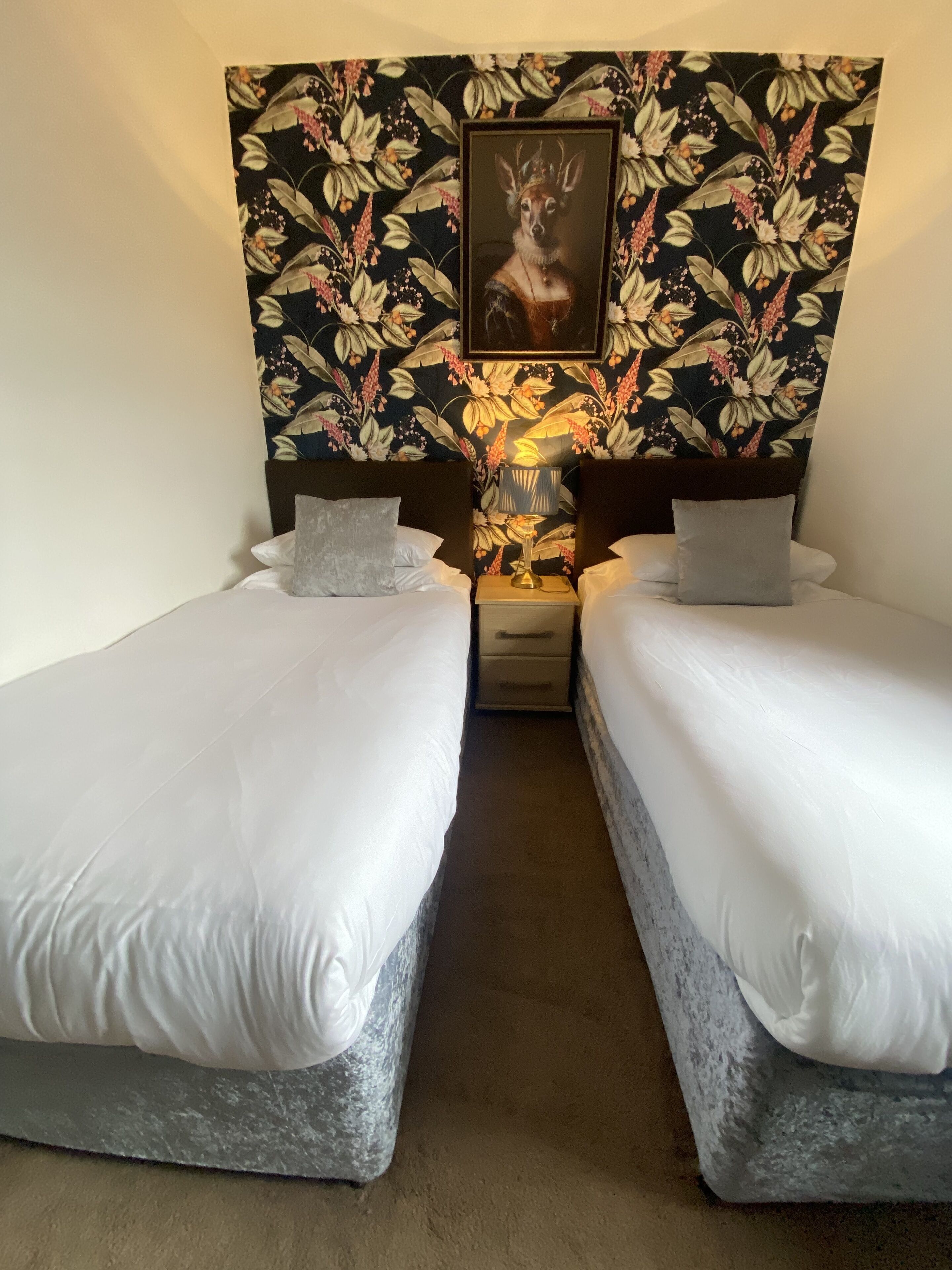 Quarto de hotel com duas camas, cabeceira estampada com flores, um quadro emoldurado e uma luminária de cabeceira.